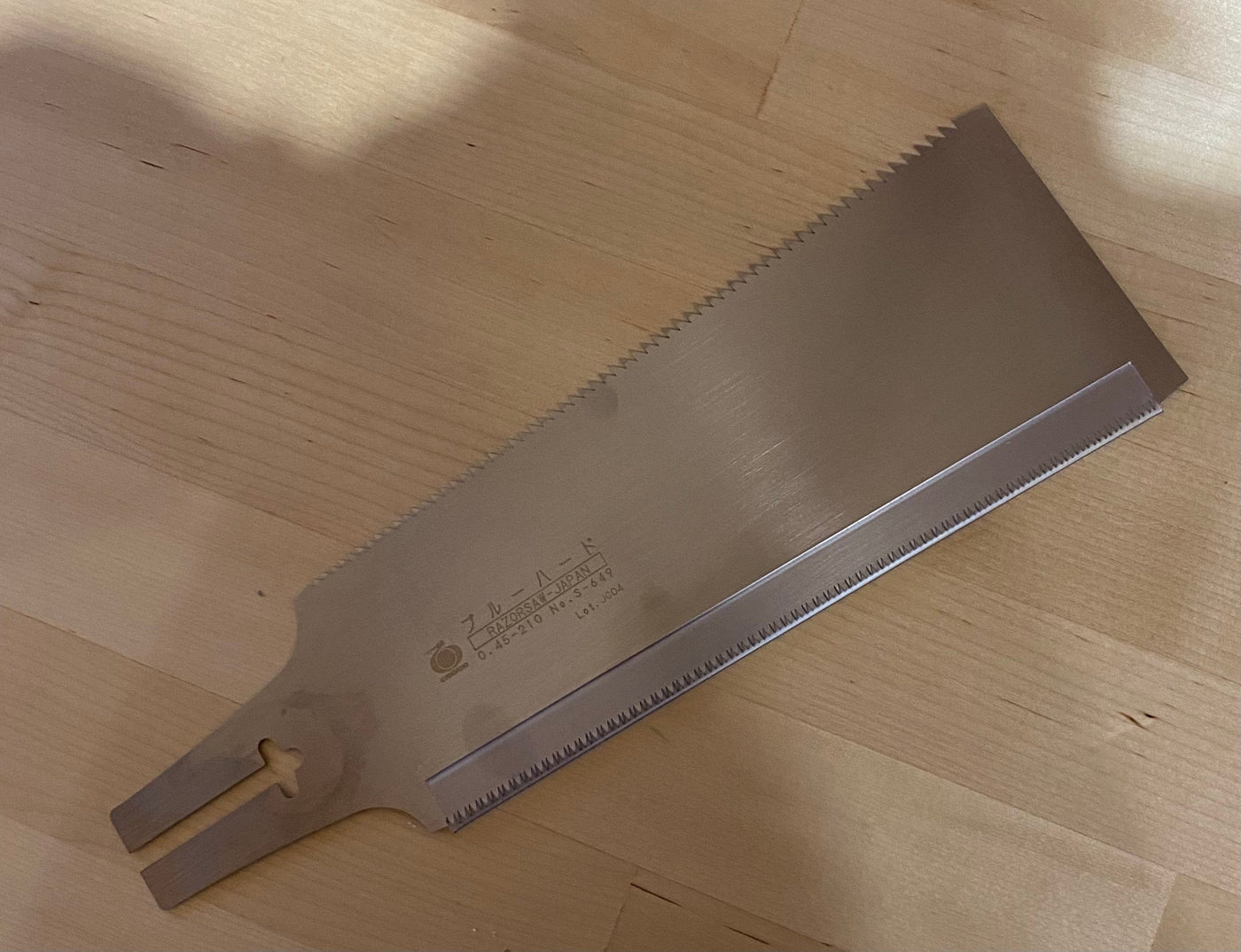 The RYOBA BLUE HARD saw by BBT, featuring a metal blade with rip cut teeth, rests on a wooden surface—ideal for woodworking enthusiasts.