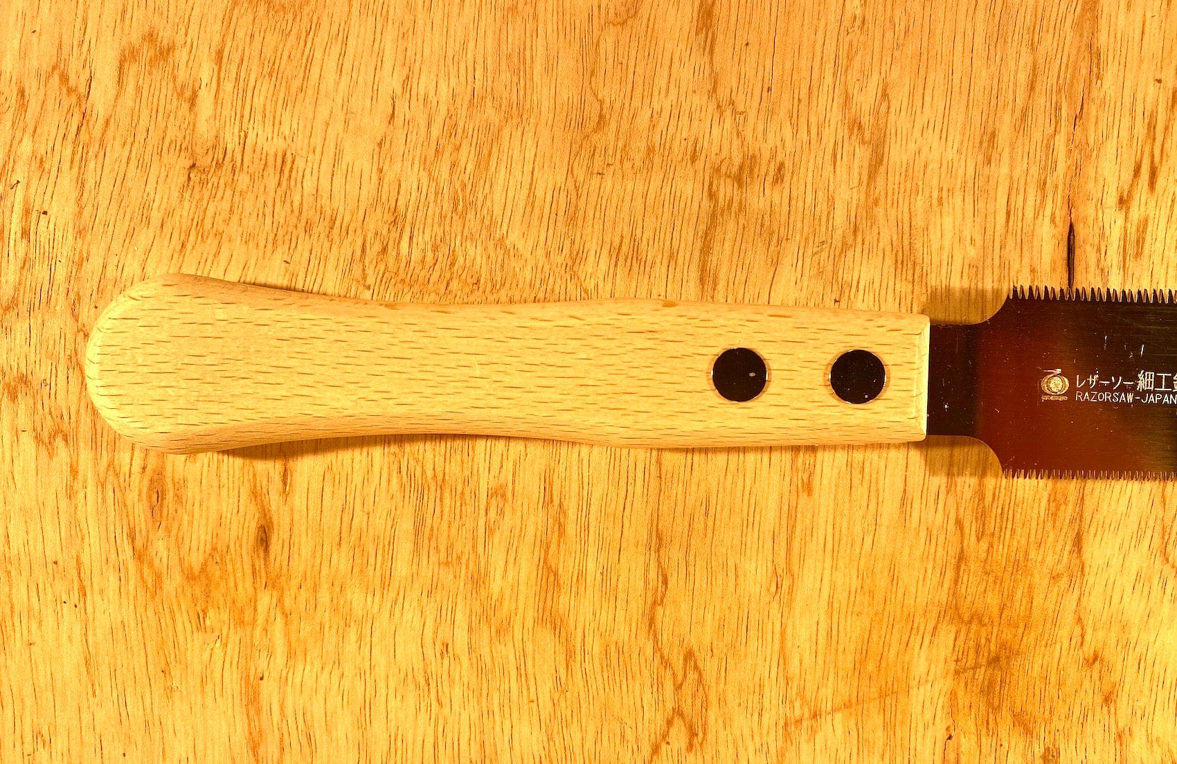 Close-up of a BBT Gyokucho Flush Cutting Saw Double Edge with a beech wood handle lying on a wooden surface, showing two black rivets securing the handle and featuring sharp cross-cut teeth.