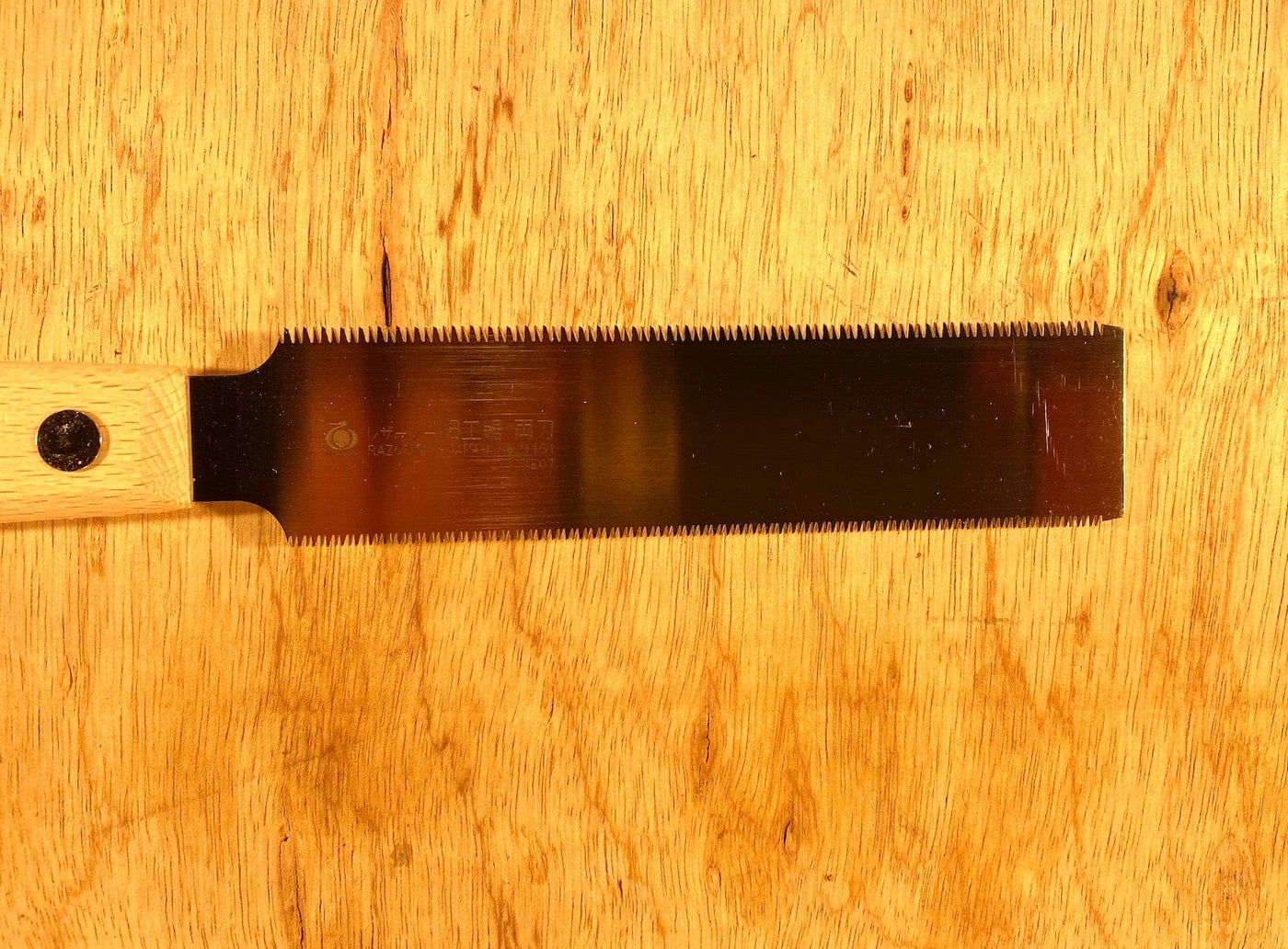 The Gyokucho Flush Cutting Saw Double Edge by BBT, featuring cross-cut teeth and a wooden handle, rests gracefully on the beech wood surface.