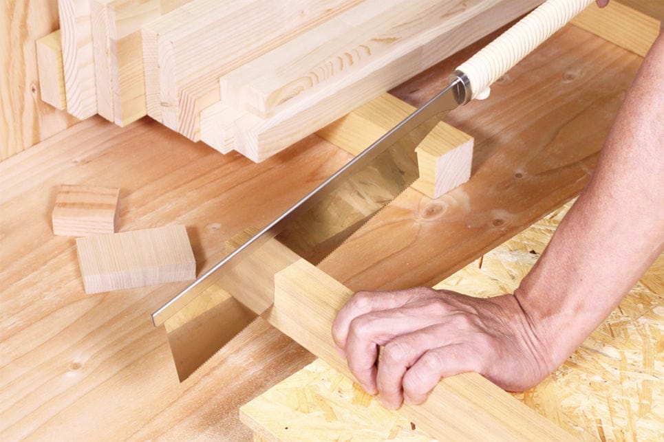 A hand is using the Razorsaw 240 For Detail Work by BBT to cut a piece of wood on a workbench, surrounded by various wooden planks and blocks, showcasing precision woodworking skills.
