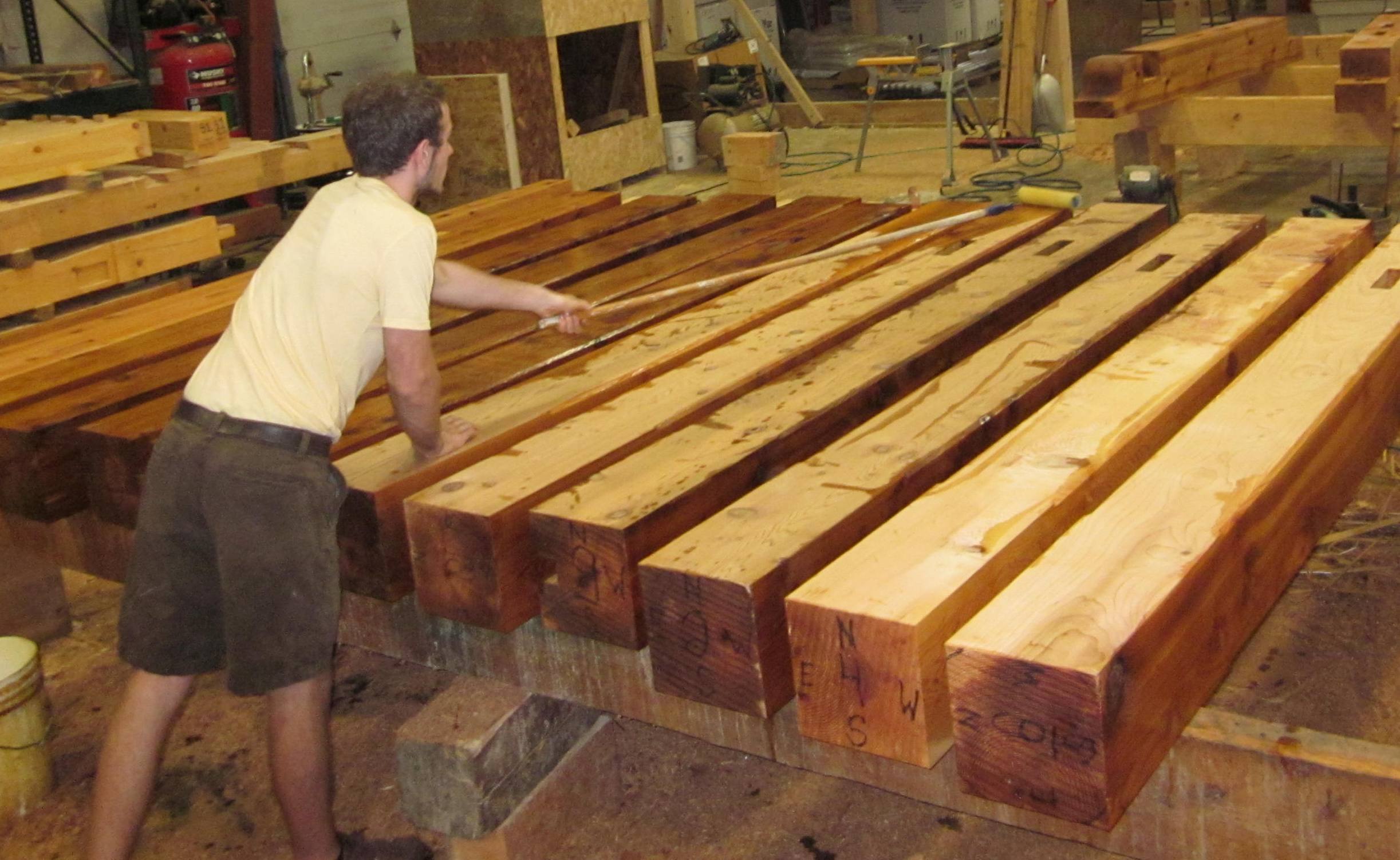 In a workshop filled with tools and materials, a man precisely measures large wooden beams to ensure accuracy for improved water resistance, using HNF's Heritage Natural Finishes Oil Select Interior & Exterior.
