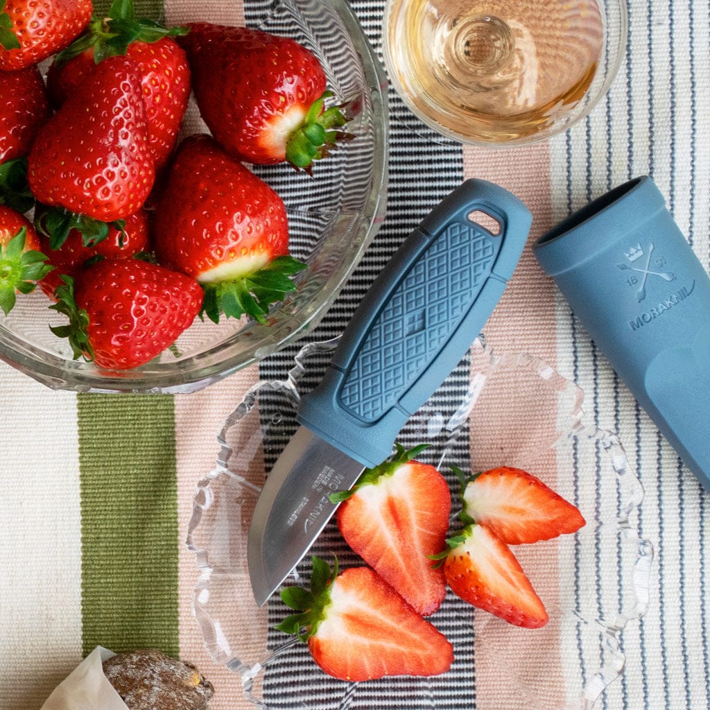 A bowl of strawberries, a Mora Eldris Light Duty knife by Industrial Revolution with a blue TPE rubber grip, sliced strawberries on a glass plate, and a wine glass sit on a striped tablecloth. The Swedish stainless steel blade gleams in the light.