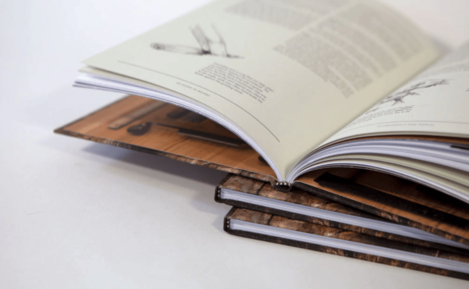 An open "Slöjd in Wood" book by LAP, with technical drawings and text, rests elegantly on two closed wooden-covered books. This scene reflects the craftsmanship reminiscent of Jögge Sundqvist's woodworking mastery, all set on a pristine white surface.