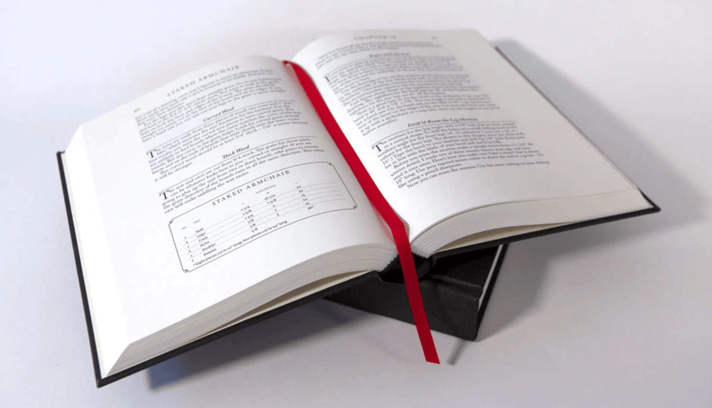 An open hardcover copy of LAP’s The Anarchist’s Design Book: Expanded Edition, with white pages and a red ribbon bookmark, sits on a closed black box on a white surface—ideal for woodworking and timeless furniture design enthusiasts.