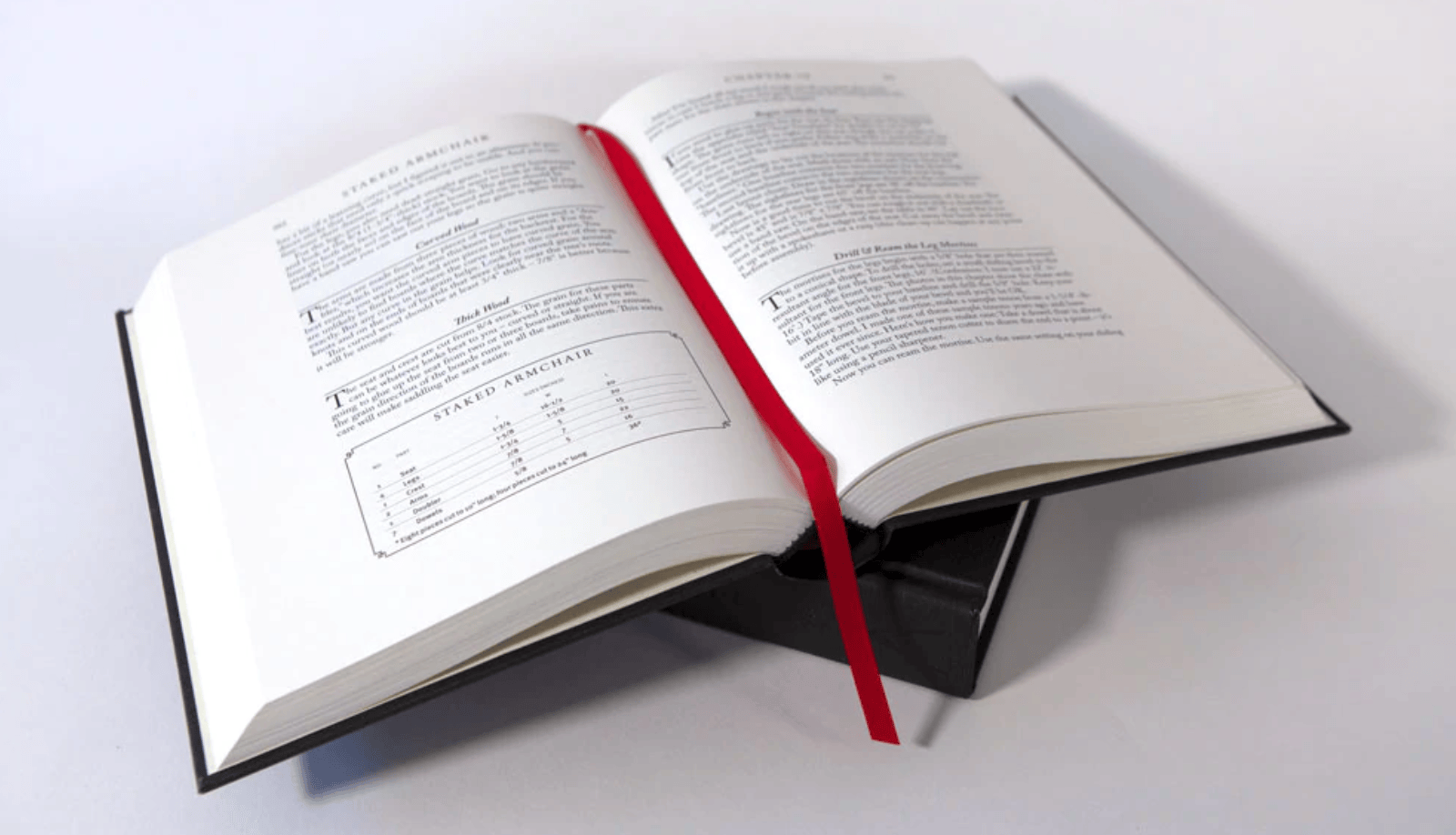 An open hardcover copy of LAP’s The Anarchist’s Design Book: Expanded Edition, with white pages and a red ribbon bookmark, sits on a closed black box on a white surface—ideal for woodworking and timeless furniture design enthusiasts.