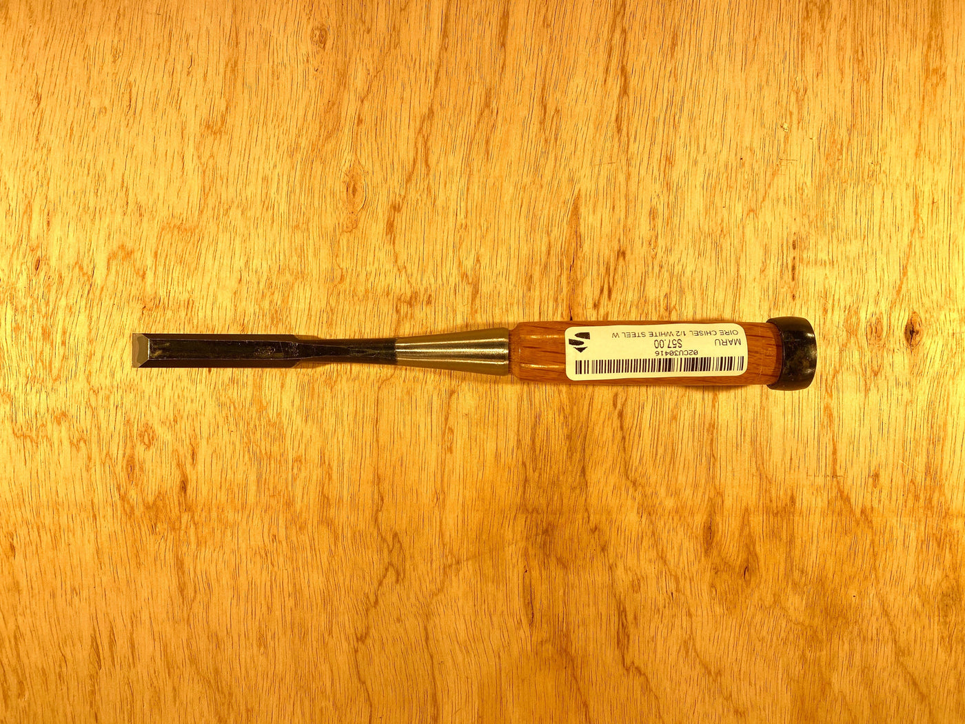 A MARU Fujikawa Oire Bench Chisel, featuring a black handle and metal blade marked "1/2 inch," lies on a wooden surface. Crafted from white steel laminated for durability, this professional-grade tool ensures precision in every cut and includes a barcode.