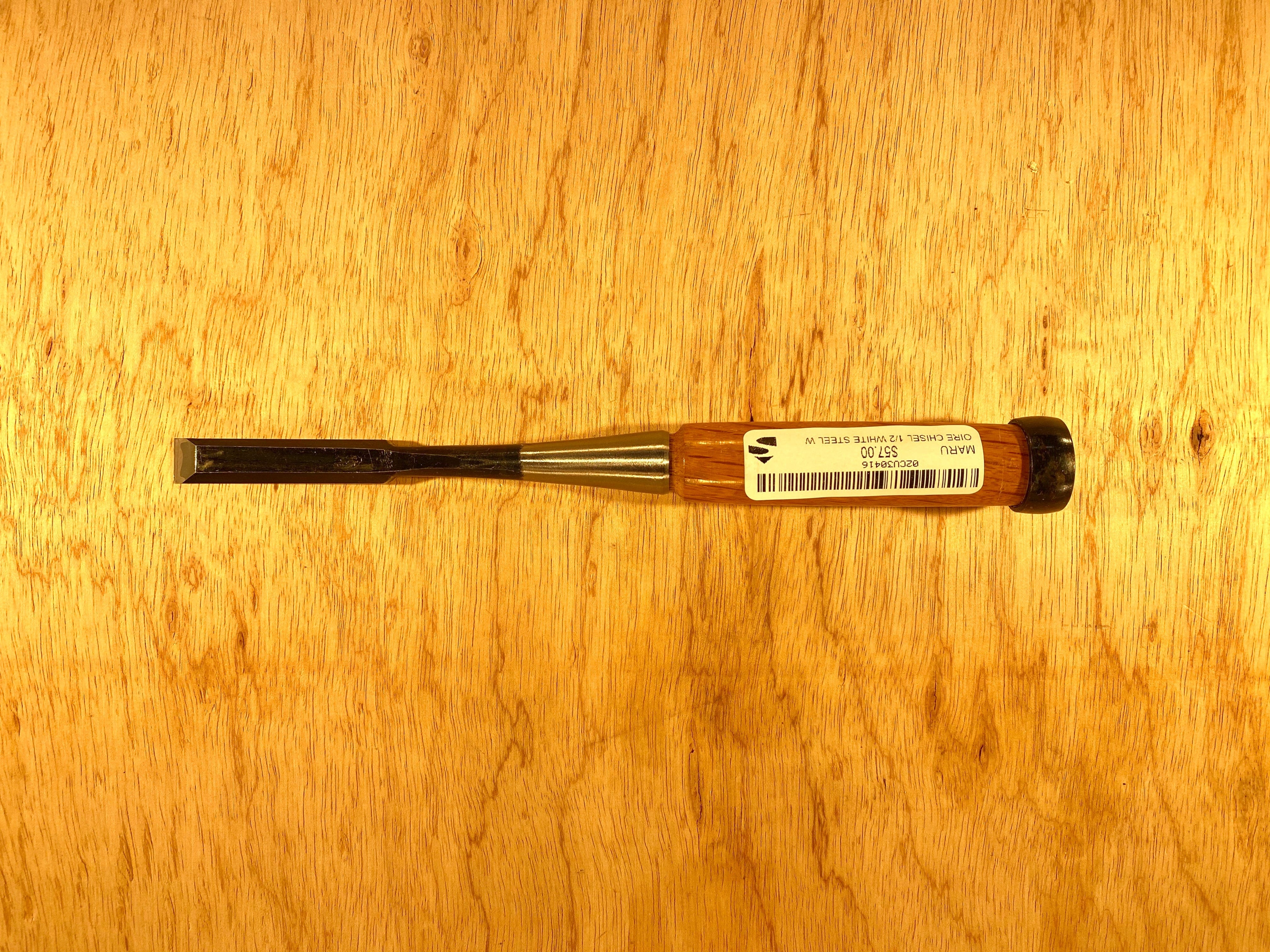 A MARU Fujikawa Oire Bench Chisel, featuring a black handle and metal blade marked "1/2 inch," lies on a wooden surface. Crafted from white steel laminated for durability, this professional-grade tool ensures precision in every cut and includes a barcode.