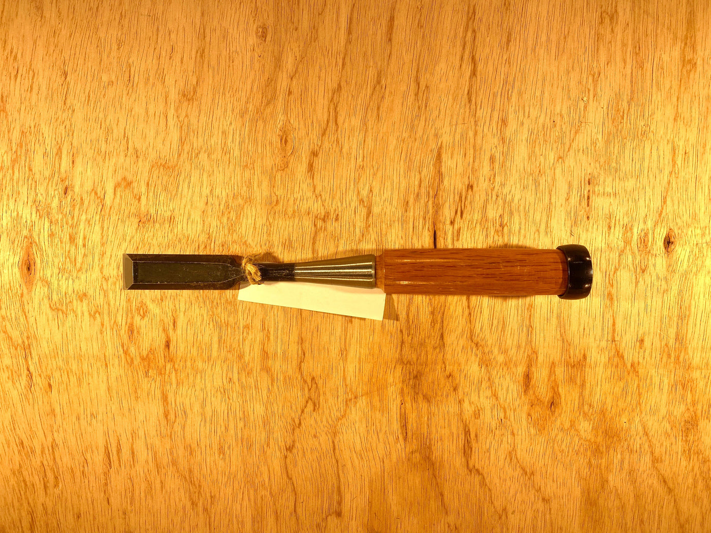 The Fujikawa Oire Bench Chisel by MARU, featuring a wooden handle and a laminated white steel blade, rests elegantly on a wood surface, exemplifying the sophistication of Japanese chisels. A small white tag is attached to the handle.