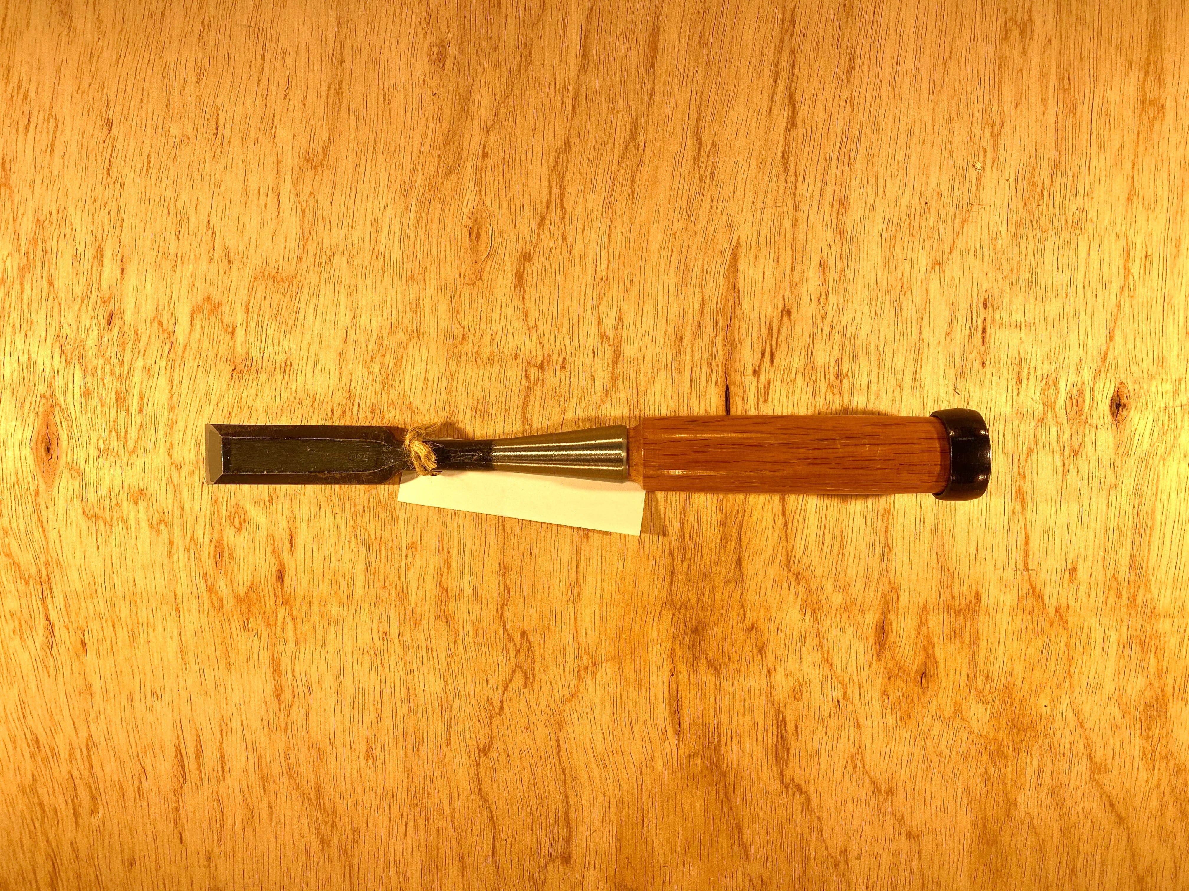 The Fujikawa Oire Bench Chisel by MARU, featuring a wooden handle and a laminated white steel blade, rests elegantly on a wood surface, exemplifying the sophistication of Japanese chisels. A small white tag is attached to the handle.