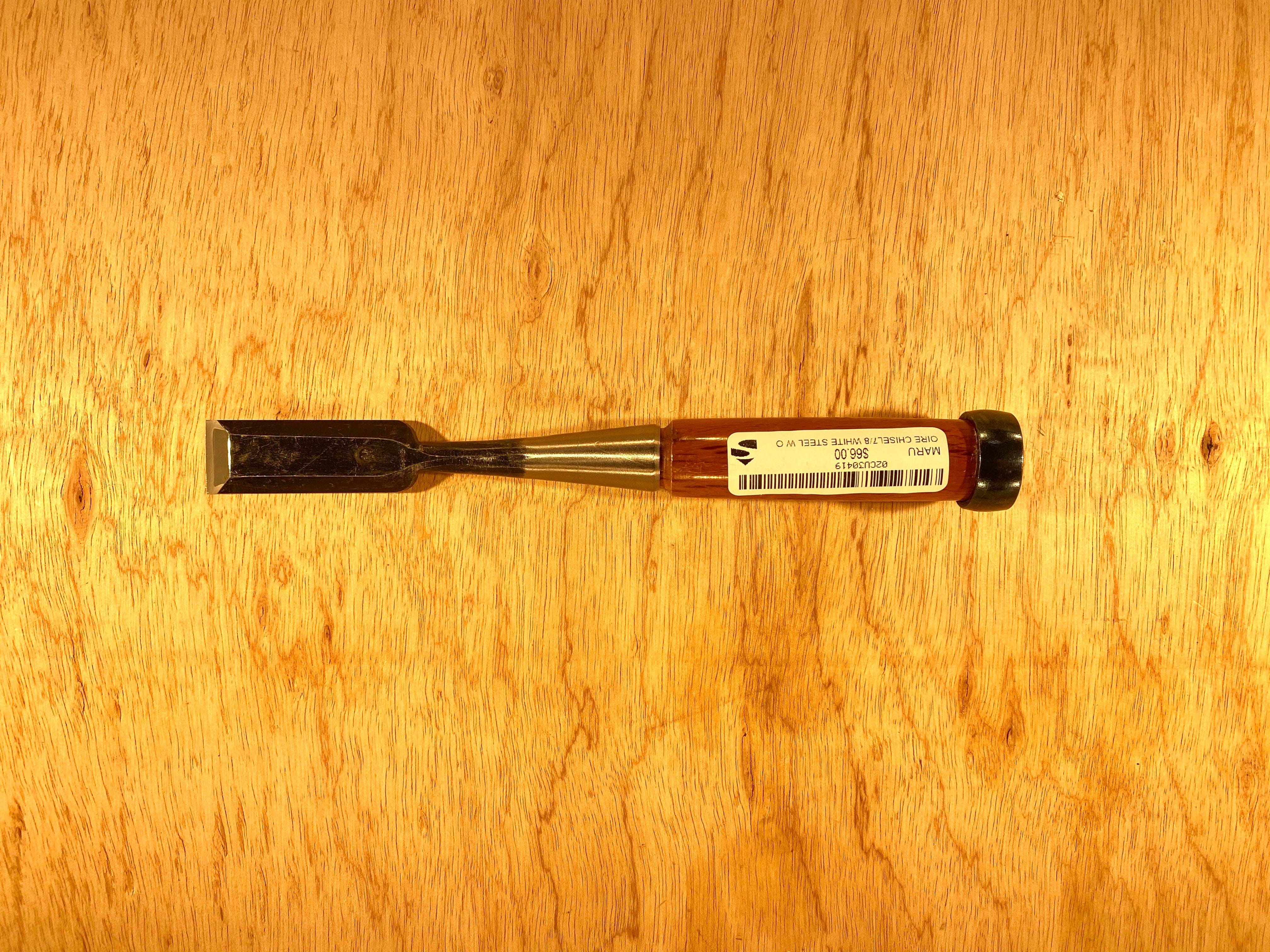 The MARU Fujikawa Oire Bench Chisel, a professional-grade tool with a white steel laminated blade and a stylish black and brown handle, rests elegantly on a wooden surface.