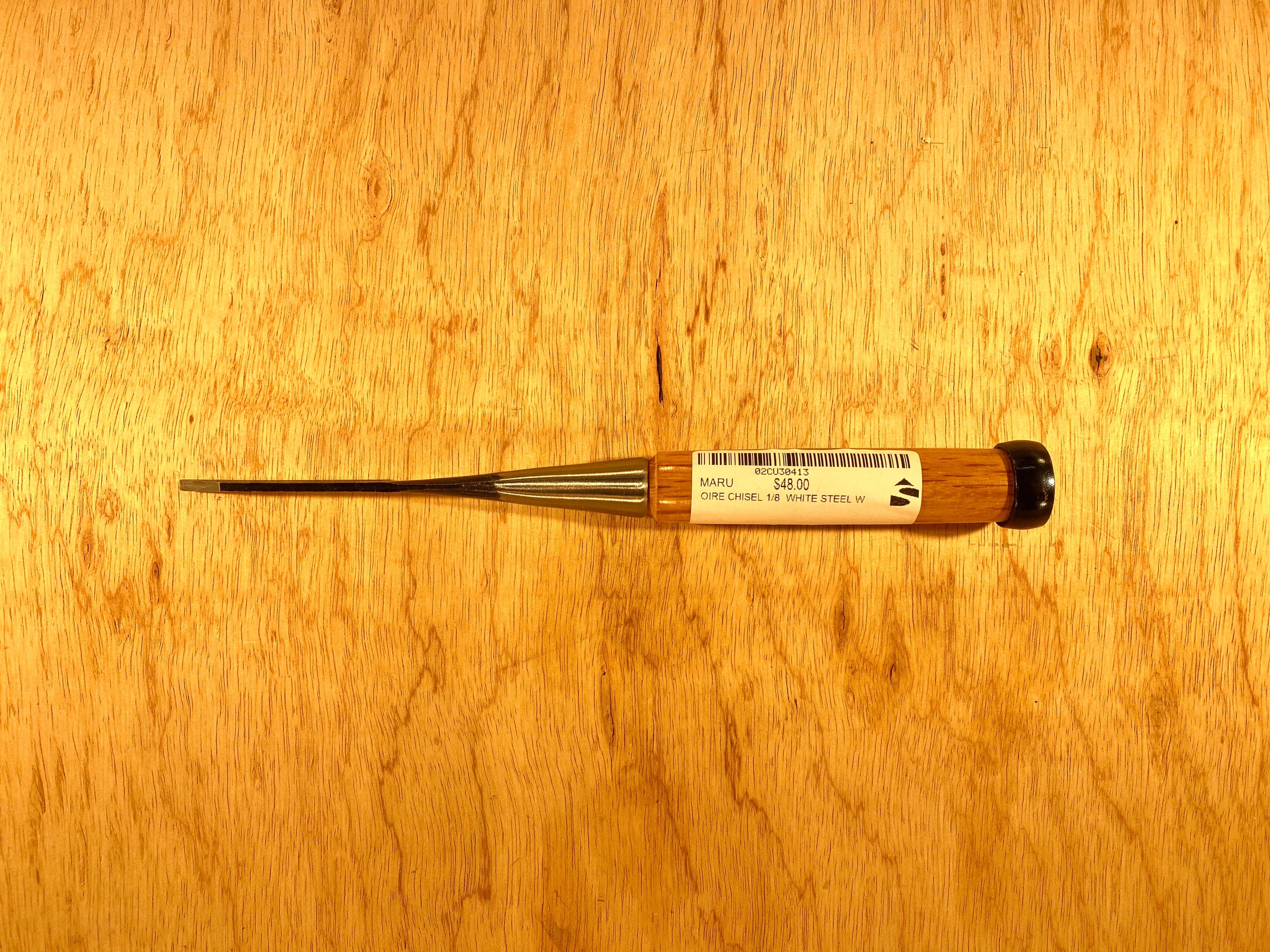 A professional-grade Fujikawa Oire Bench Chisel by MARU with a wooden handle and metal blade rests on the wooden surface. A label with text adorns the handle, showcasing its craftsmanship. Ideal for precision work, this tool embodies the artistry of Japanese chisels crafted from white steel laminated for durability.