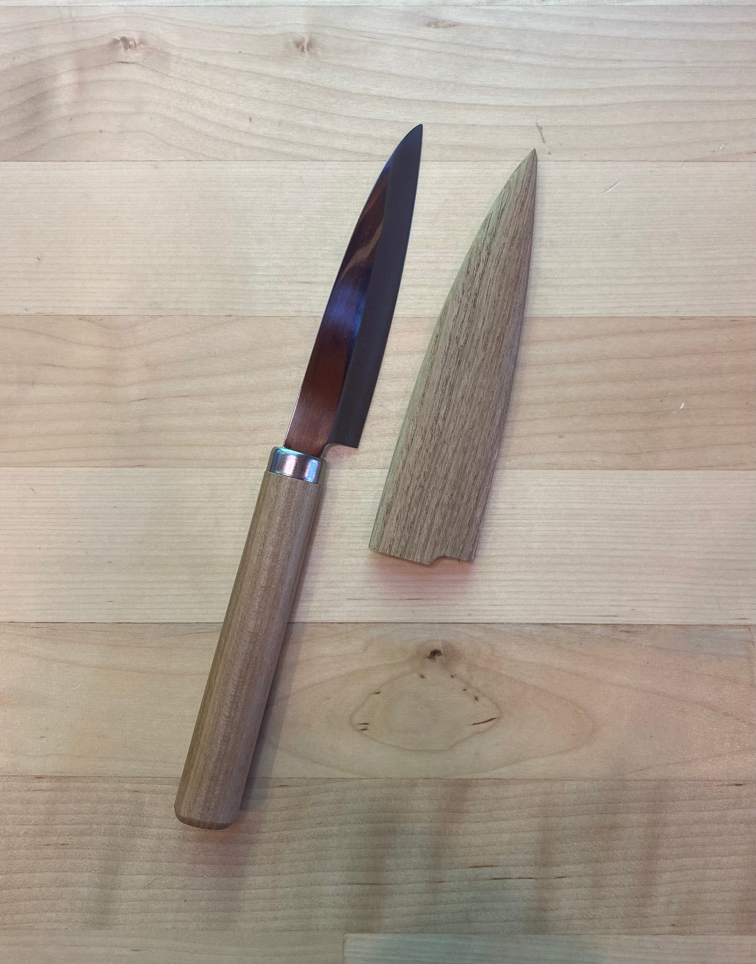 A small MARU FRUIT KNIFE with an oak handle and blade cover lies on a wooden surface. The stainless steel blade is partially covered by the sheath, which is placed next to it.