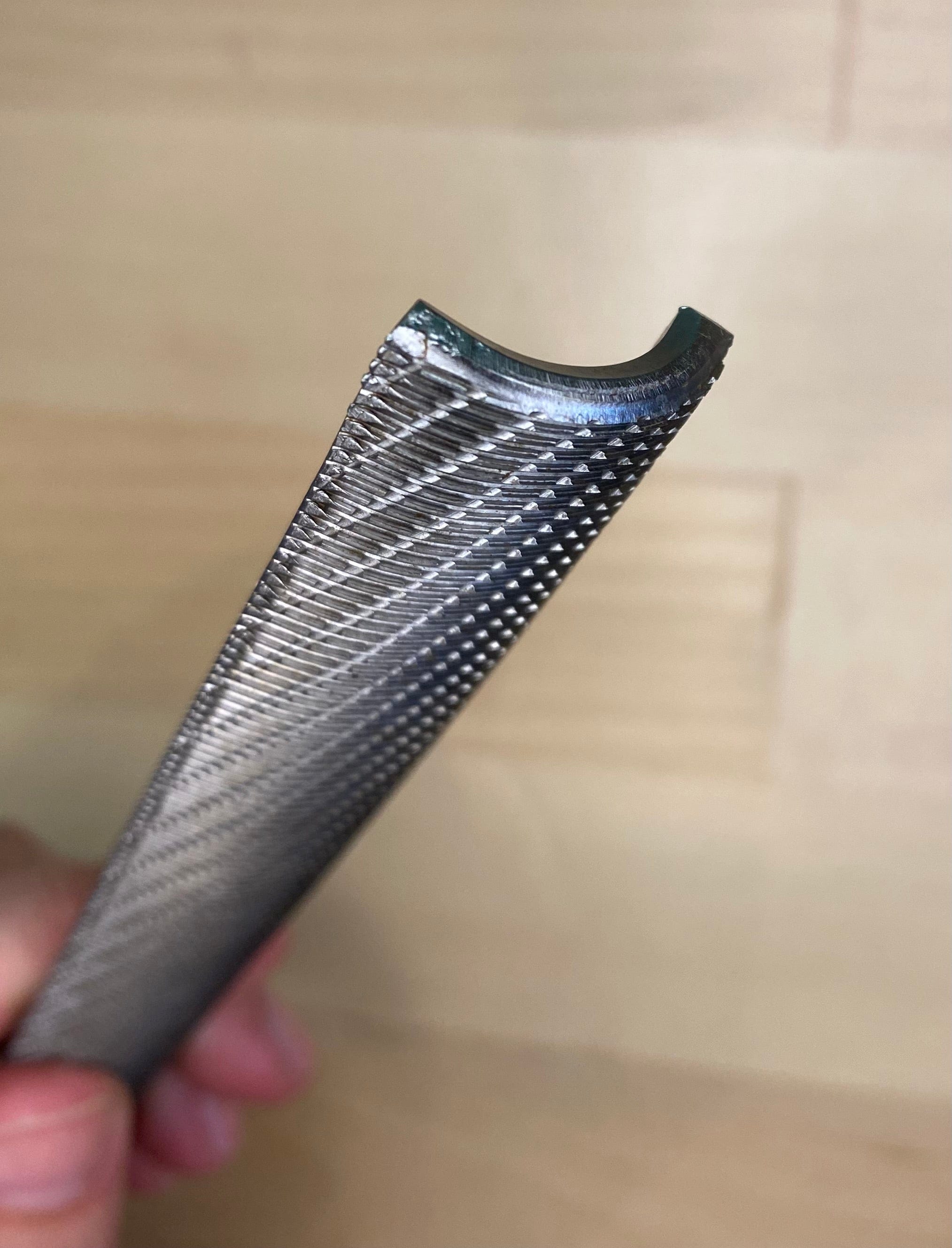 A close-up of a curved MARU Iwasaki Wood File, featuring a diamond-patterned textured surface known for its fast cutting action, held in a person's hand against a wooden backdrop.