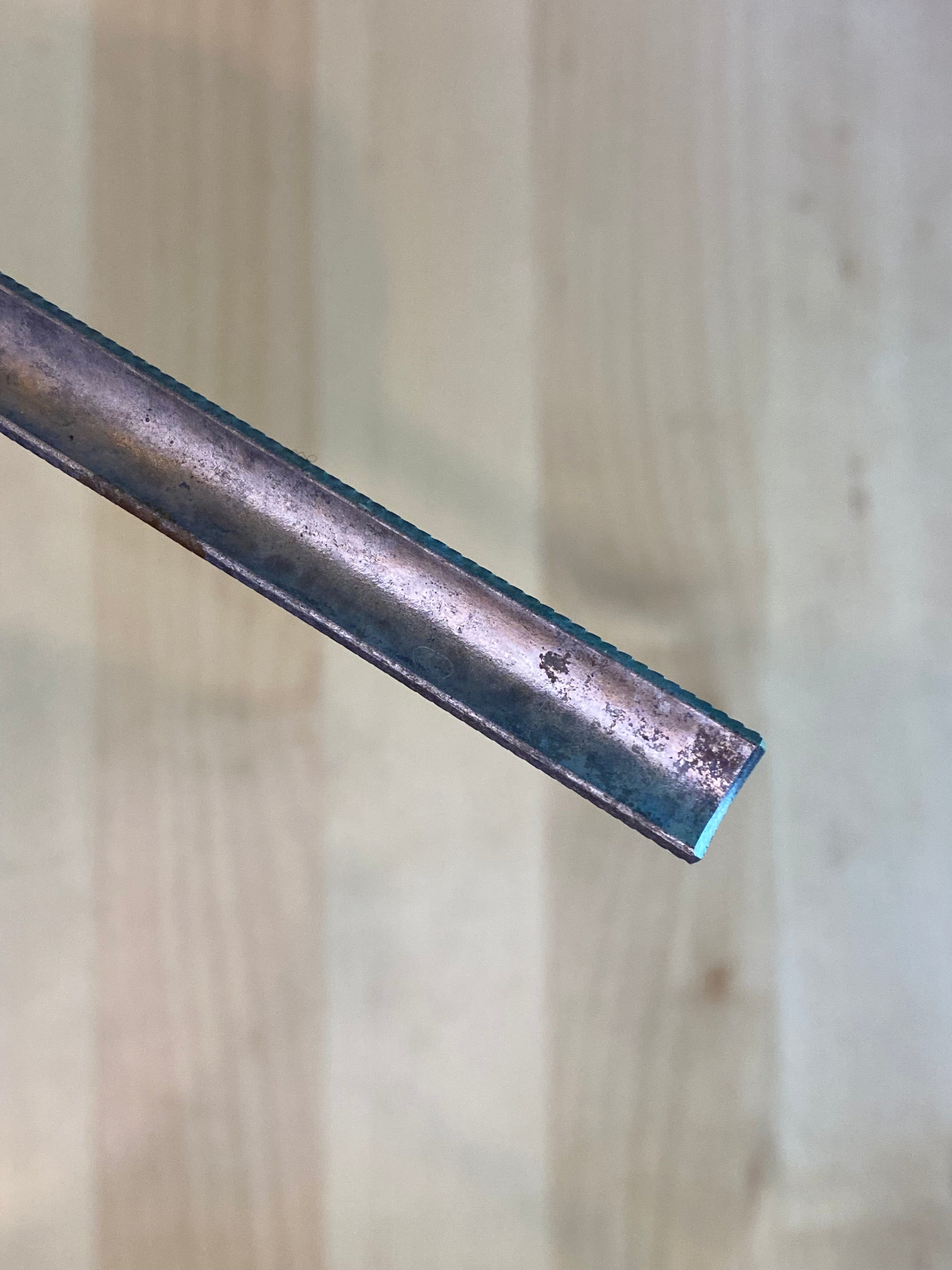 Close-up of the Iwasaki Half Round Ex Fine W/Grip by MARU, displaying a metal body with a blue interior. The weathered surface resembles a woodworker's tool, set against a light, blurred wooden backdrop that enhances its crafted ambiance.