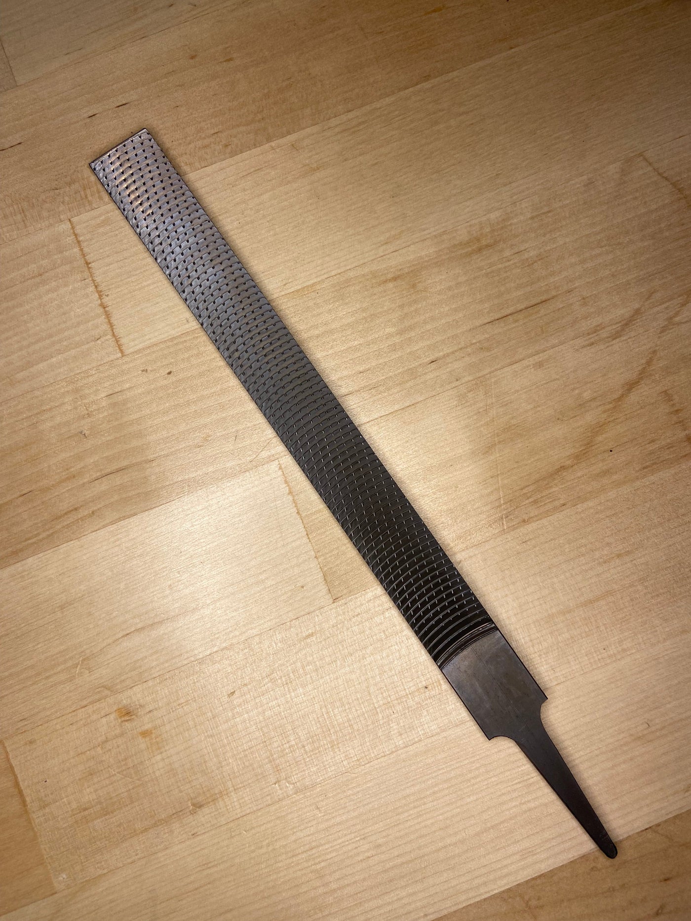 The MARU Iwasaki Flat Medium Cut Wood File, featuring a textured abrasive surface and pointed tang, rests on a light wooden surface.