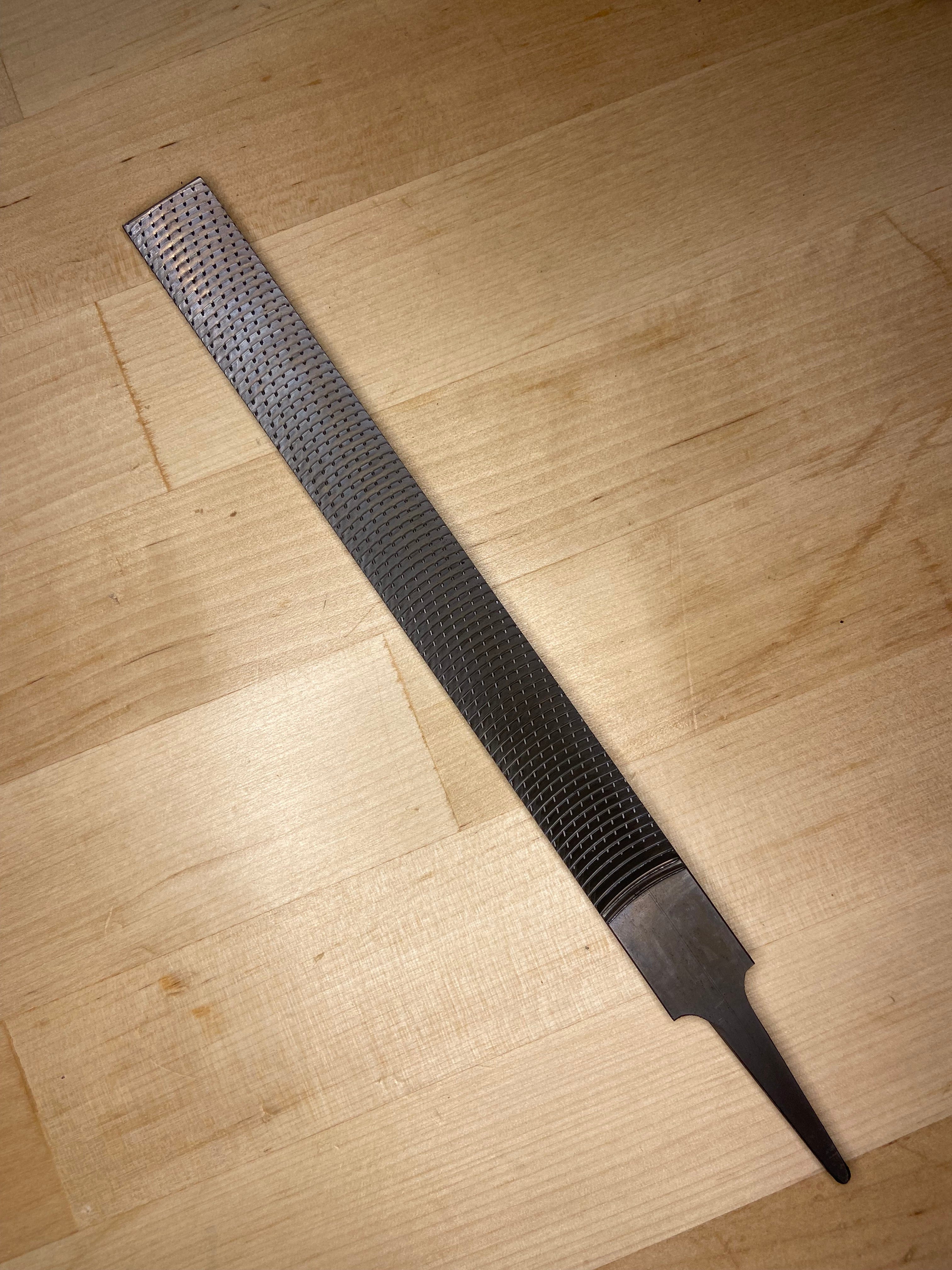 The MARU Iwasaki Flat Medium Cut Wood File, featuring a textured abrasive surface and pointed tang, rests on a light wooden surface.