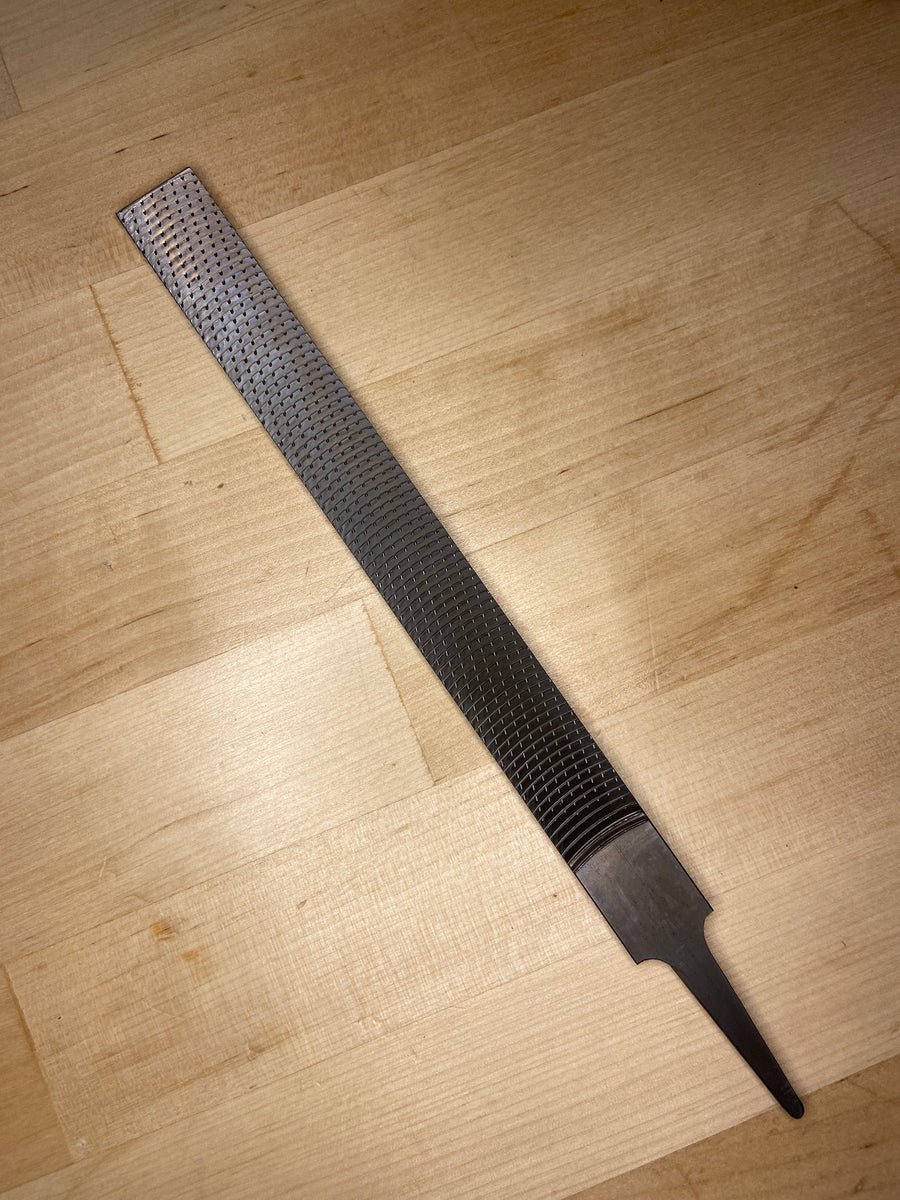 The MARU Iwasaki Flat Medium Cut Wood File, featuring a textured abrasive surface and pointed tang, rests on a light wooden surface.