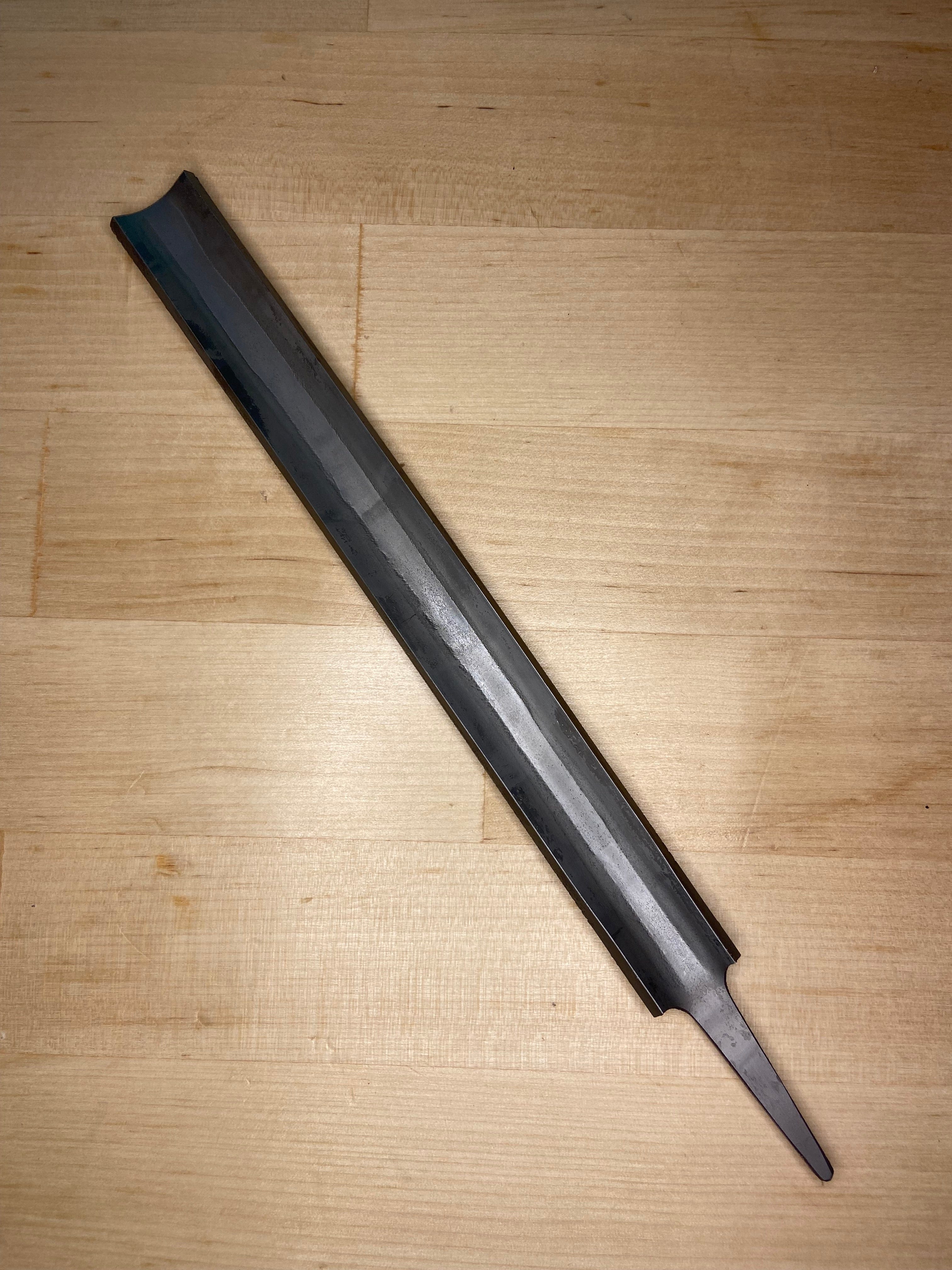 The MARU Iwasaki Half Round Medium Cut Wood File, a dark metal tool with a central ridge and pointed tip, lies on a light wooden surface. The file has no handle and appears unfinished.