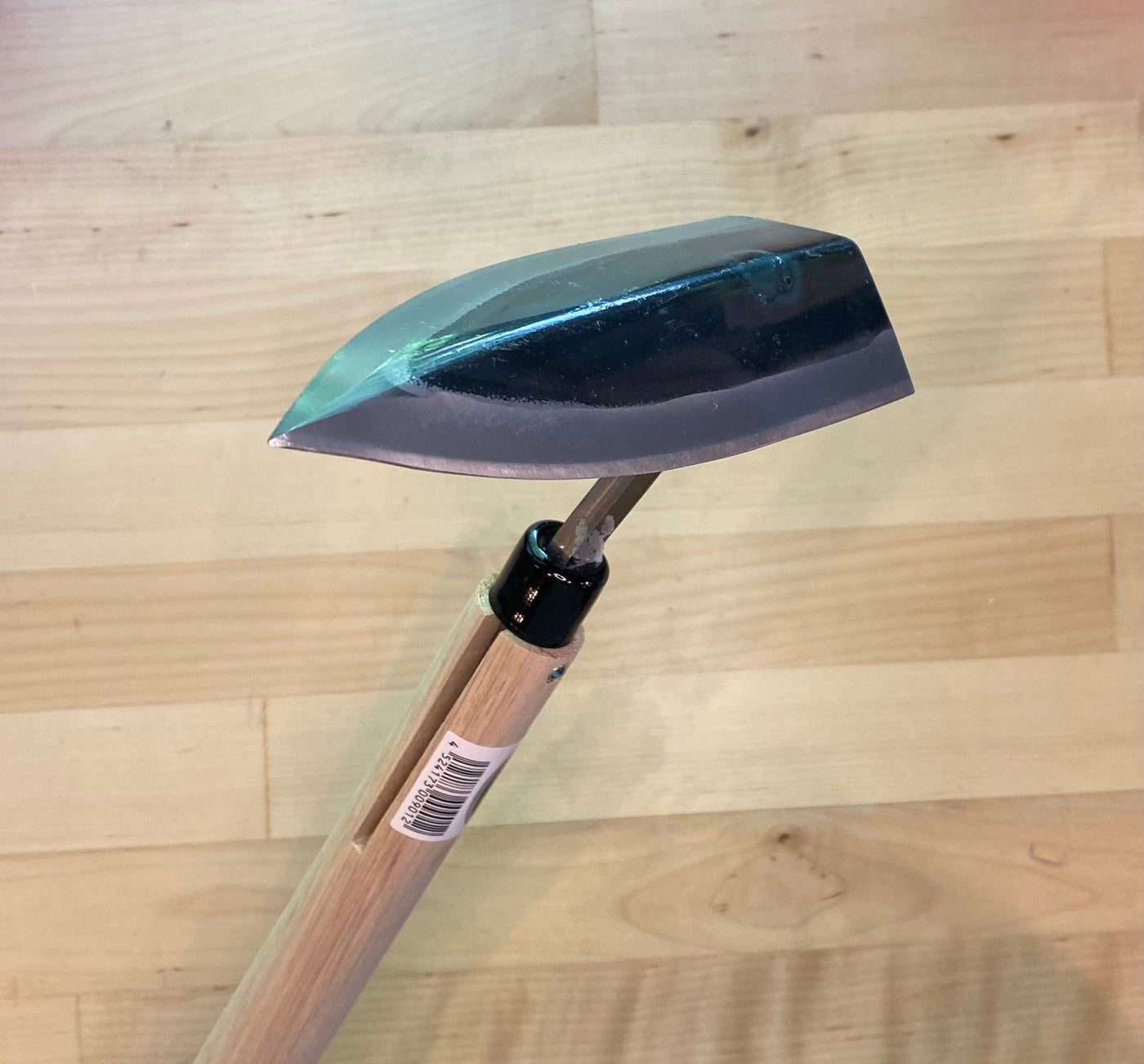 A MARU Triangle Tip Weeder with a high-carbon blade, shiny metal head, and wooden handle lies on a wooden floor, ready for digging into soil.