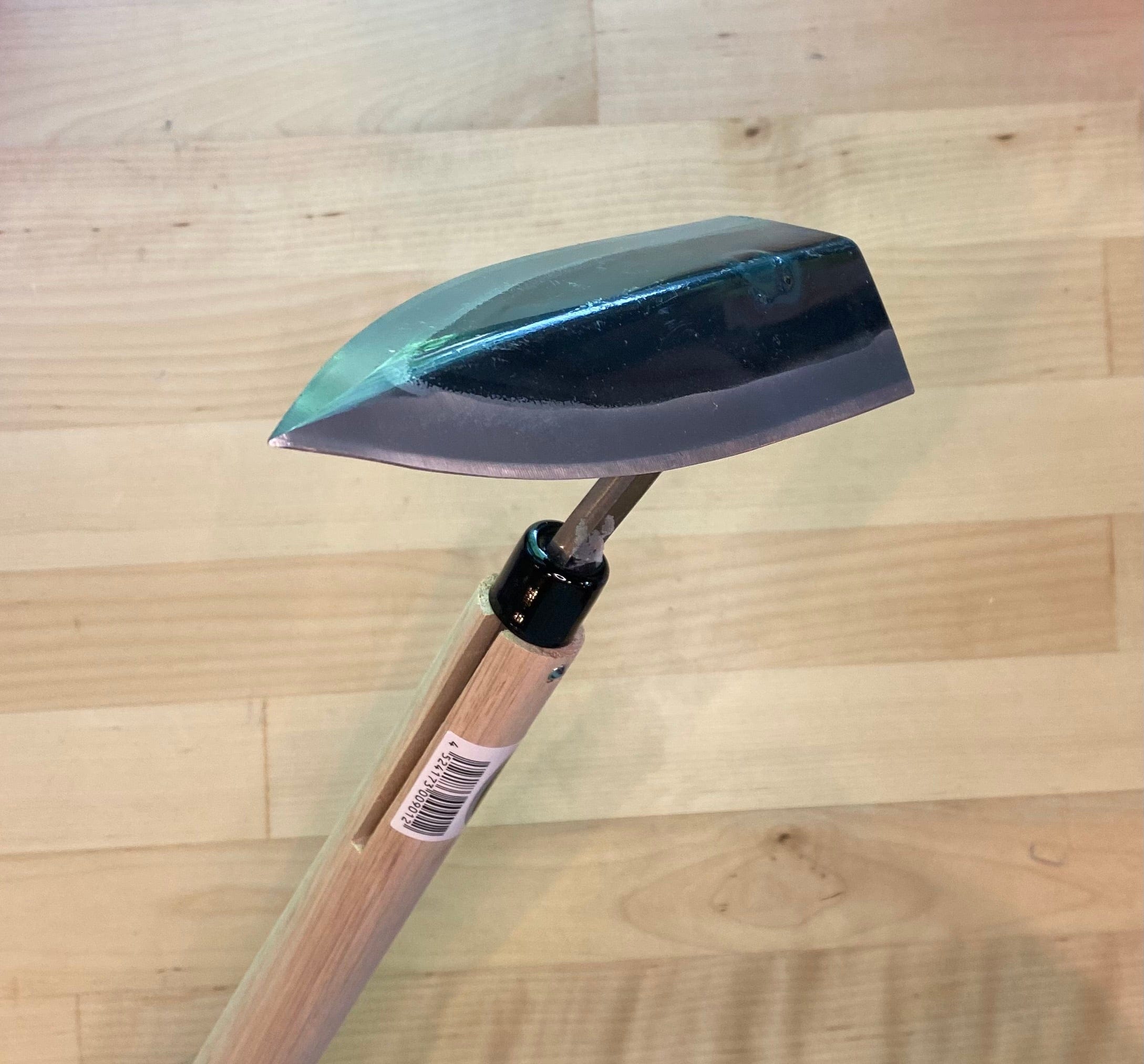 A MARU Triangle Tip Weeder with a high-carbon blade, shiny metal head, and wooden handle lies on a wooden floor, ready for digging into soil.
