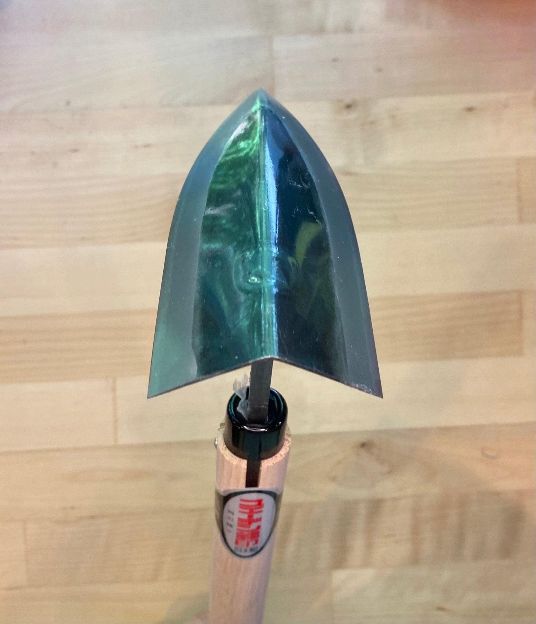 The MARU Triangle Tip Weeder features a pointed, high-carbon metal blade for efficient soil loosening, attached to a wooden handle and shown resting on a light wood surface.