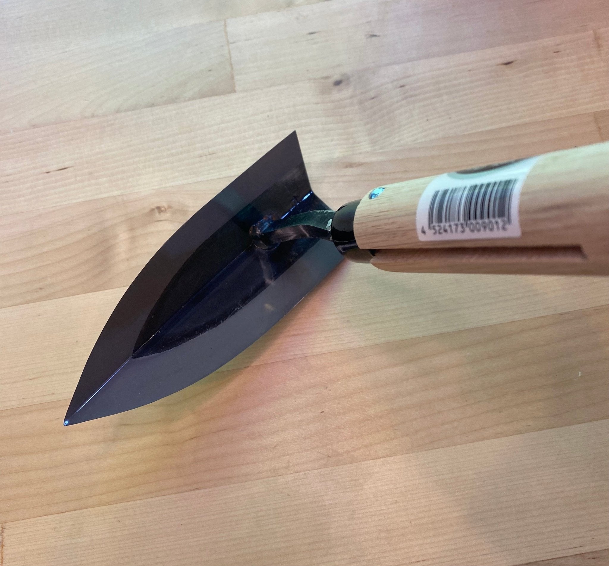 A close-up of the MARU Triangle Tip Weeder with a sharp, high-carbon blade and wooden handle, featuring a barcode sticker, resting on a light wood surface. Ideal for loosening soil in garden beds.