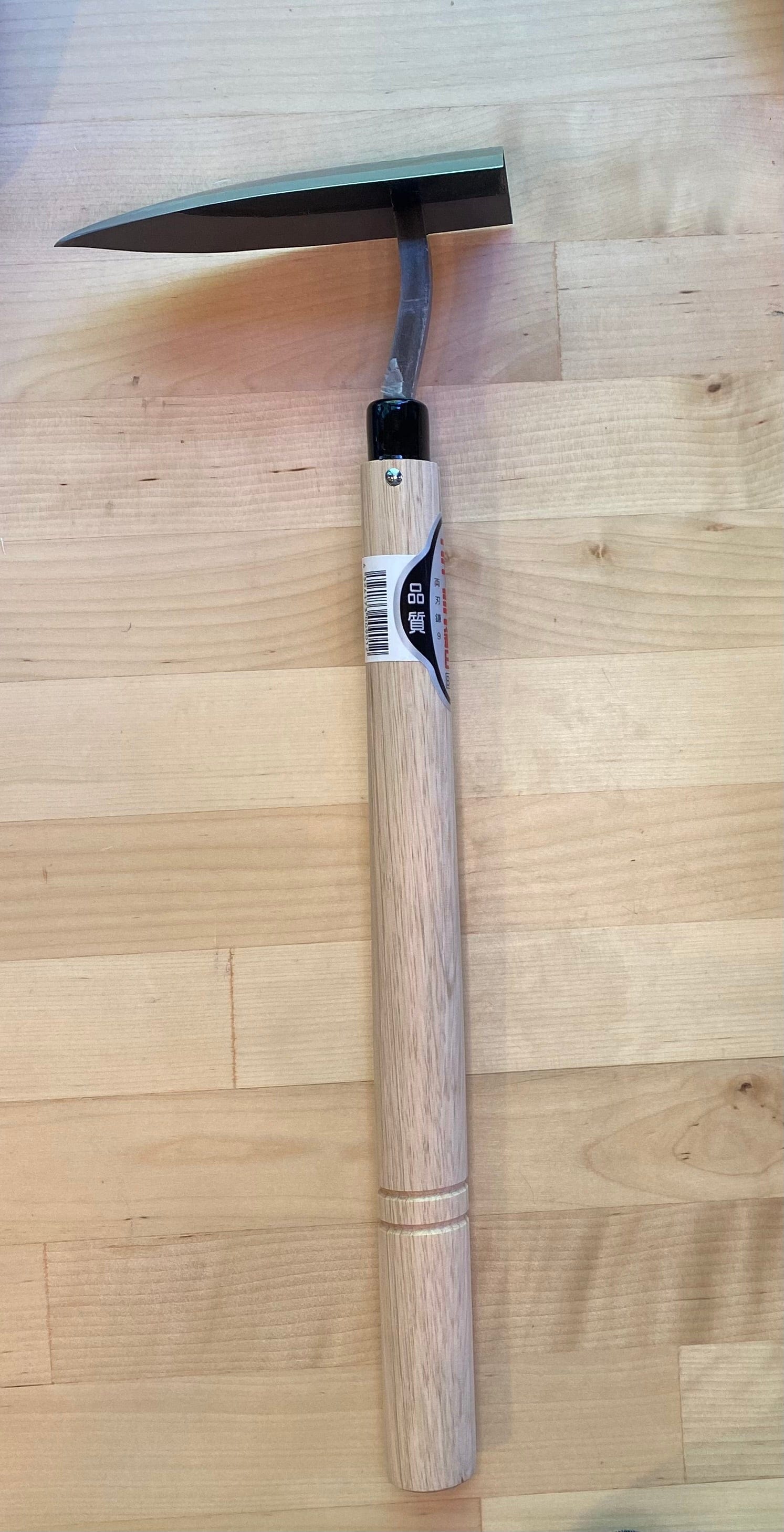 A MARU Traingle Tip Weeder with a wooden handle and a flat, high-carbon blade lies on a light wooden surface. The blade is attached at an angle, making it ideal for soil digging, and there is a barcode sticker on the handle.