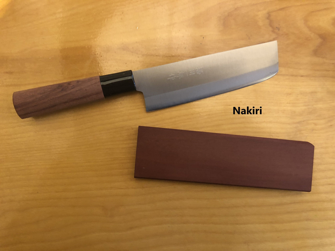 A MARU Japanese Kitchen Knife with a wooden handle and matching sheath rests on a light wood surface. The product name Nakiri is labeled next to the blade, similar to other Japanese kitchen knives.