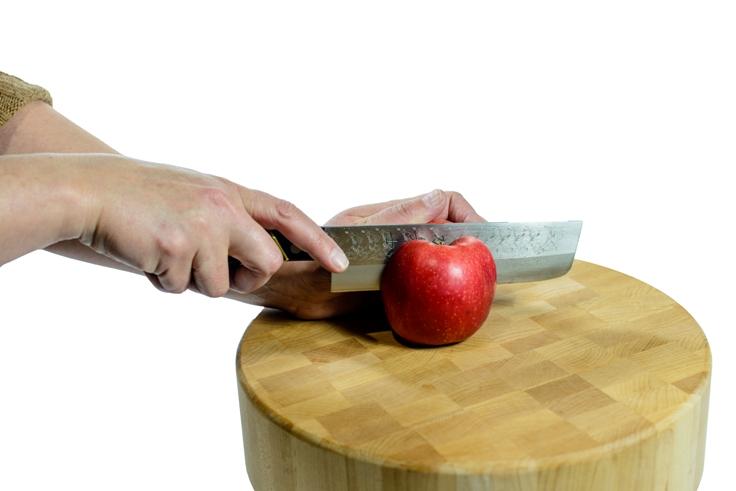 Hands are slicing a red apple using a MARU Usuba Knife on a round wooden cutting board, highlighting the knife's sleek Pakkawood handle.