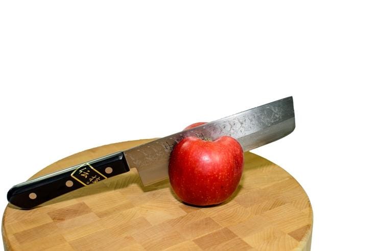 A red apple is sliced with a MARU Usuba Knife, its pakkawood handle gleaming against the plain white background, all on a wooden cutting board.
