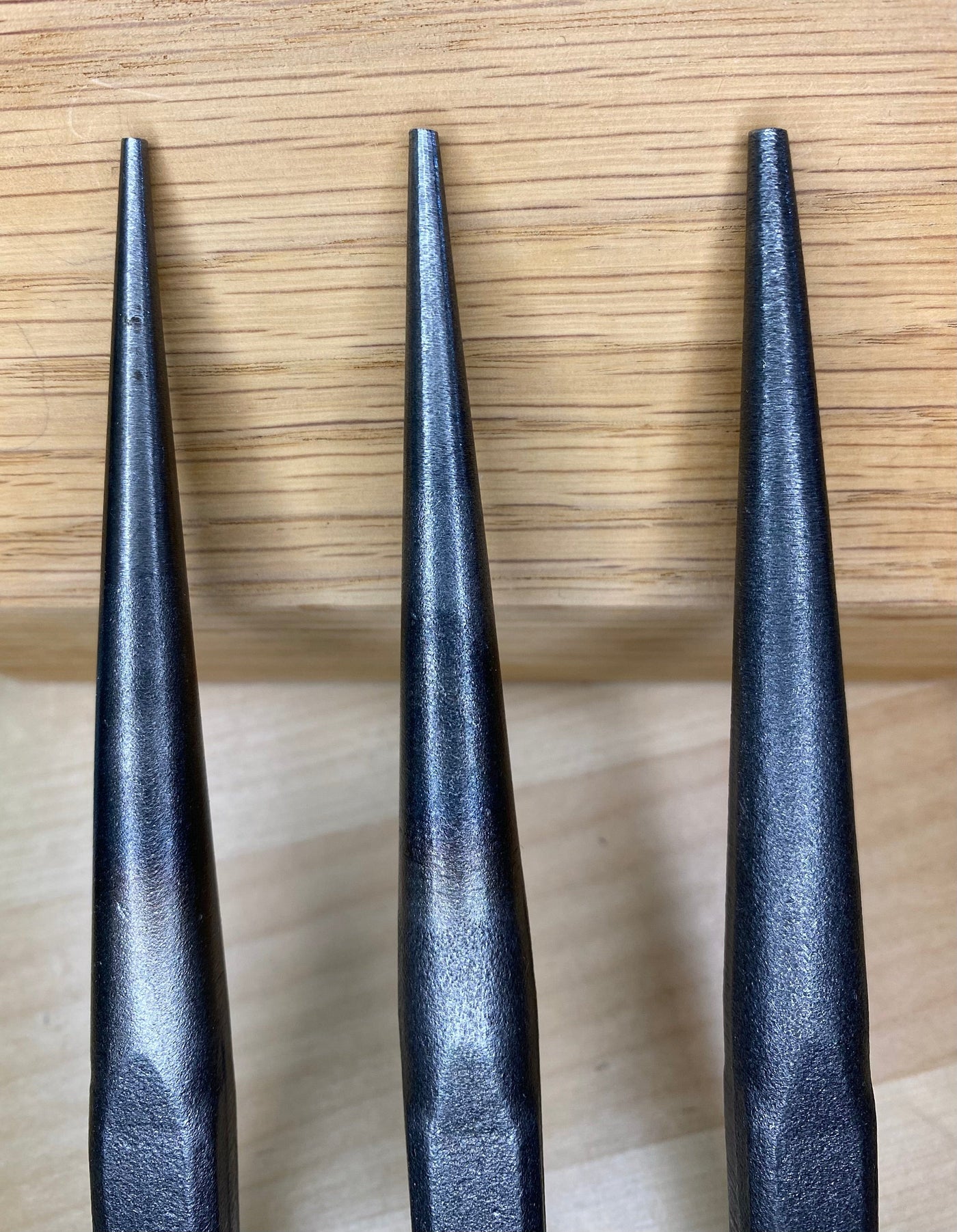 A Japanese Nail Set by MARU, featuring three black metal spikes with a design inspired by Japanese tools, stands upright on a light wooden surface against a wooden backdrop.