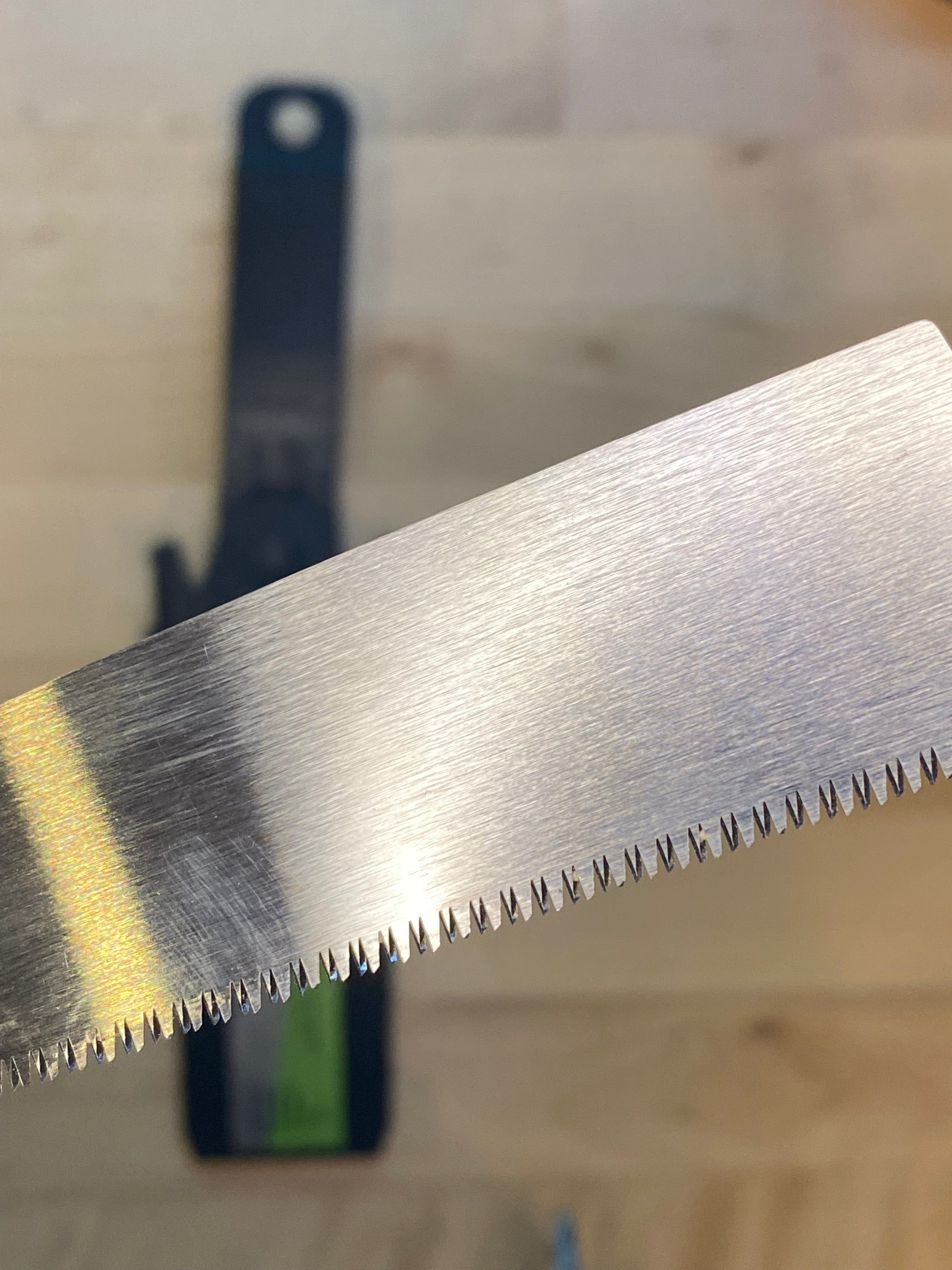 Close-up of a MARU Small Sheathed Japanese Hand Saw with small, sharp teeth, perfect for fine woodworking. A blurred tool is visible in the background. The precision of Japanese craftsmanship shines through this impulse-hardened blade.