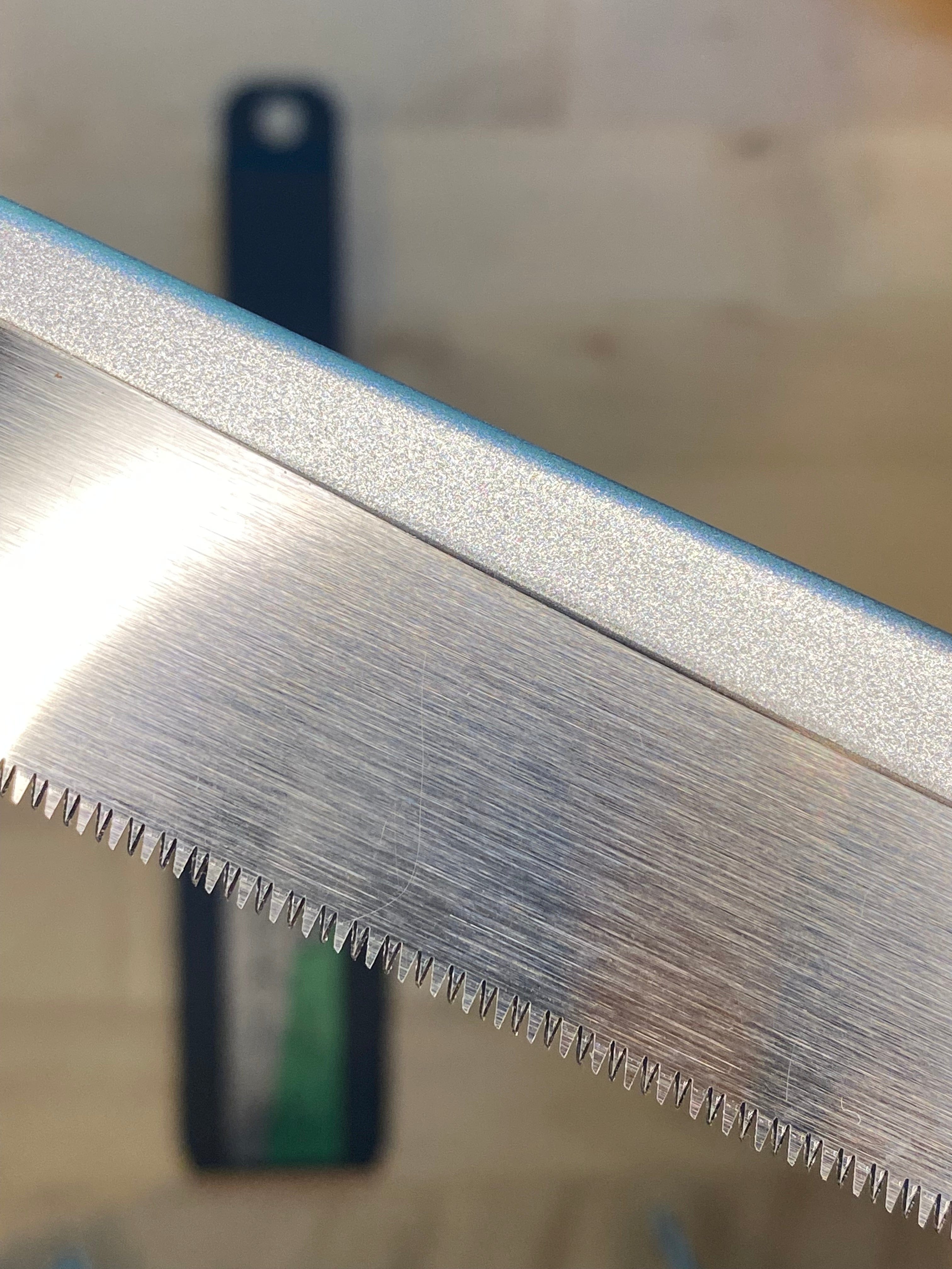 Close-up of MARU's Small Sheathed Japanese Hand Saws' impulse-hardened blades and sharp teeth. The background shows a blurry surface, possibly a tabletop, highlighting the precision ideal for fine woodworking.