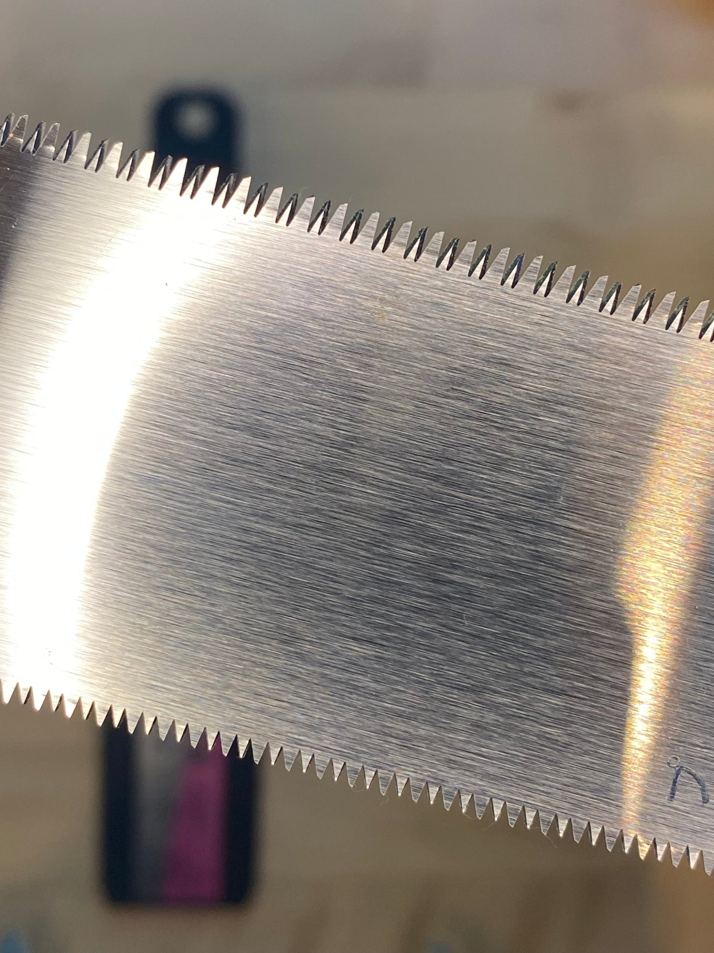 Close-up image of MARU's Small Sheathed Japanese Hand Saw, featuring a shiny, impulse-hardened serrated metal blade viewed from its side against a blurred background.