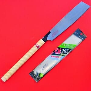 The Curved-Edge Z-Saw 265 by MARU, with its beige wrapped handle and packaging, displayed against a bright red background. This 10 ½" joinery saw features a sharp blade with impulse-hardened teeth for superior durability.