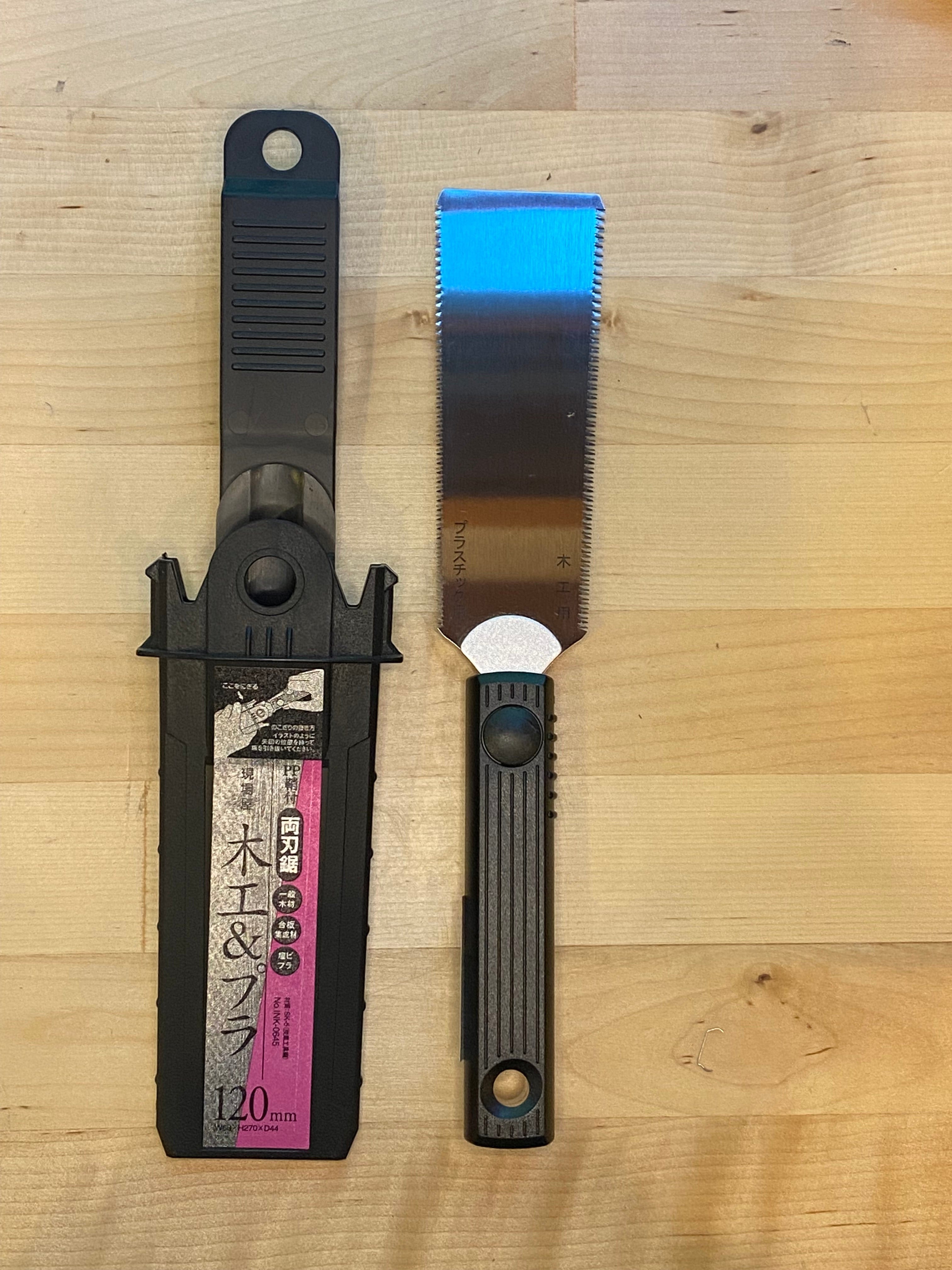 A Small Sheathed Japanese Hand Saw by MARU, featuring a black handle and an impulse-hardened, serrated blade, is placed beside its black plastic sheath on a wooden surface.