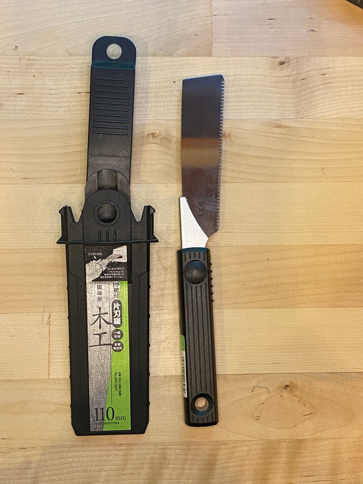 The MARU Small Sheathed Japanese Hand Saw, with a distinctive black and green handle and impulse-hardened blades, is displayed alongside its black plastic sheath on a wooden surface.