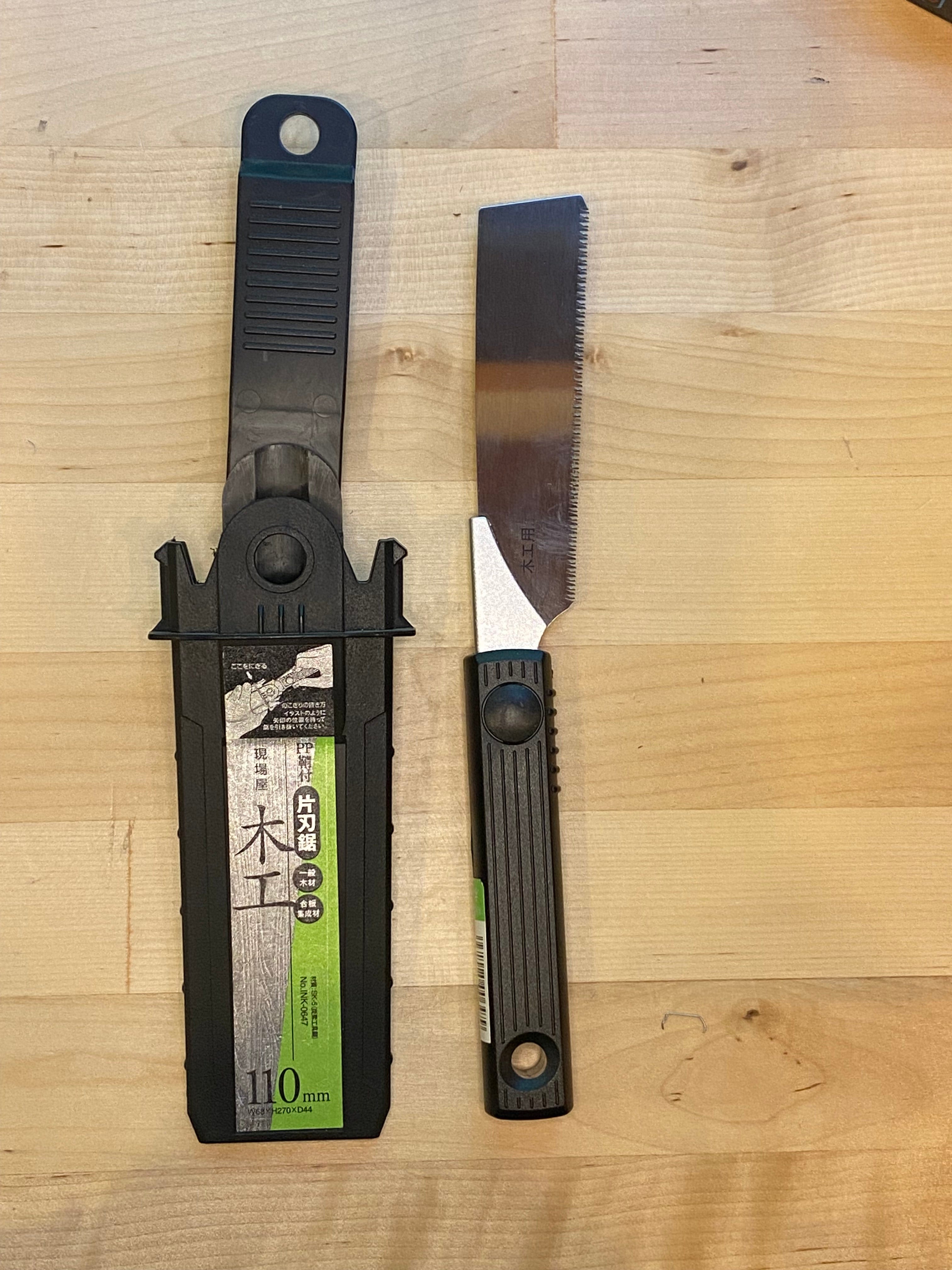 The MARU Small Sheathed Japanese Hand Saw, with a distinctive black and green handle and impulse-hardened blades, is displayed alongside its black plastic sheath on a wooden surface.