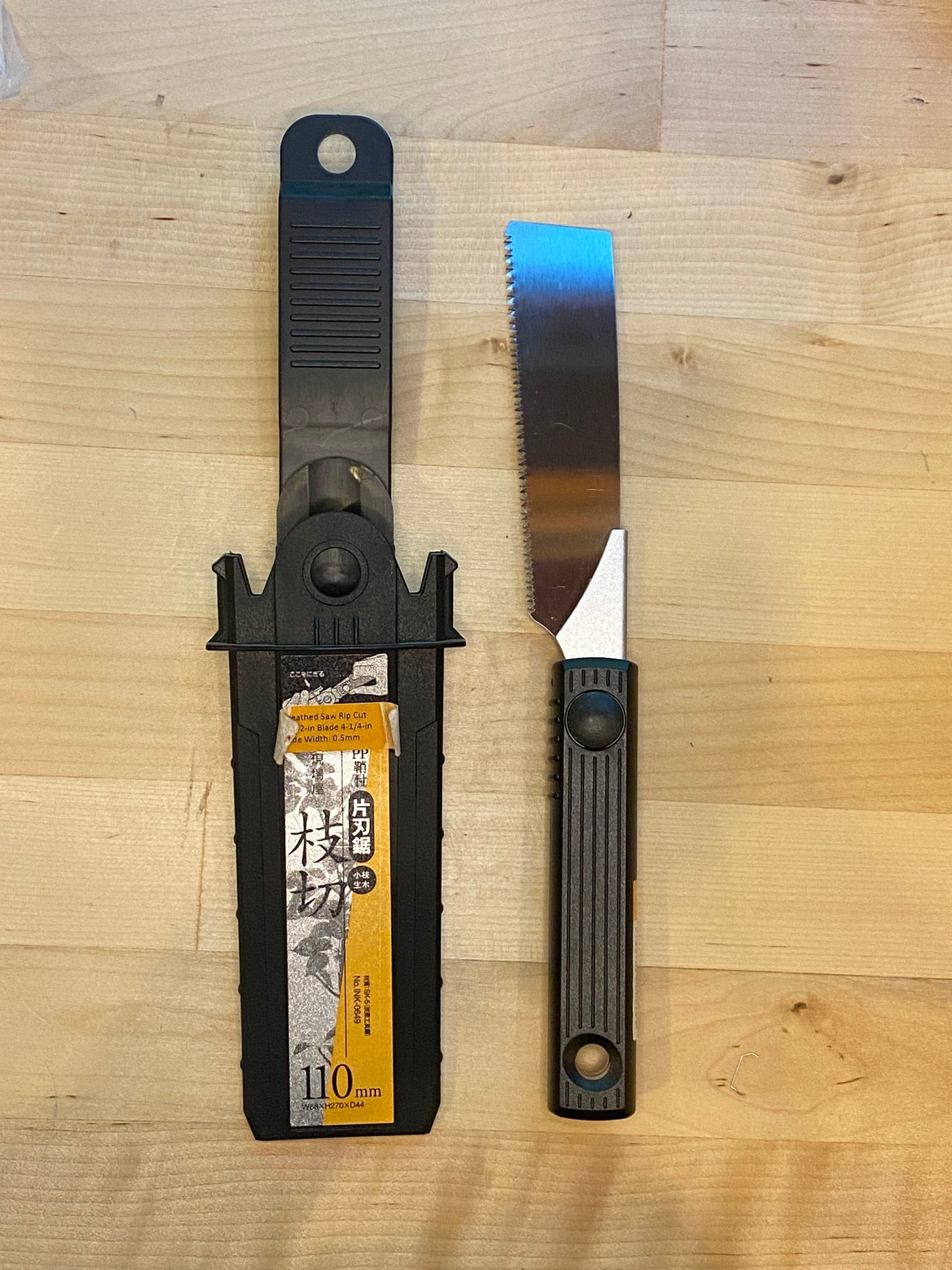 A MARU Small Sheathed Japanese Hand Saw with a black and silver handle lies next to its black plastic sheath, which features a label with text and illustrations, on a wooden surface. This fine woodworking tool boasts an impulse-hardened blade that is partially serrated.