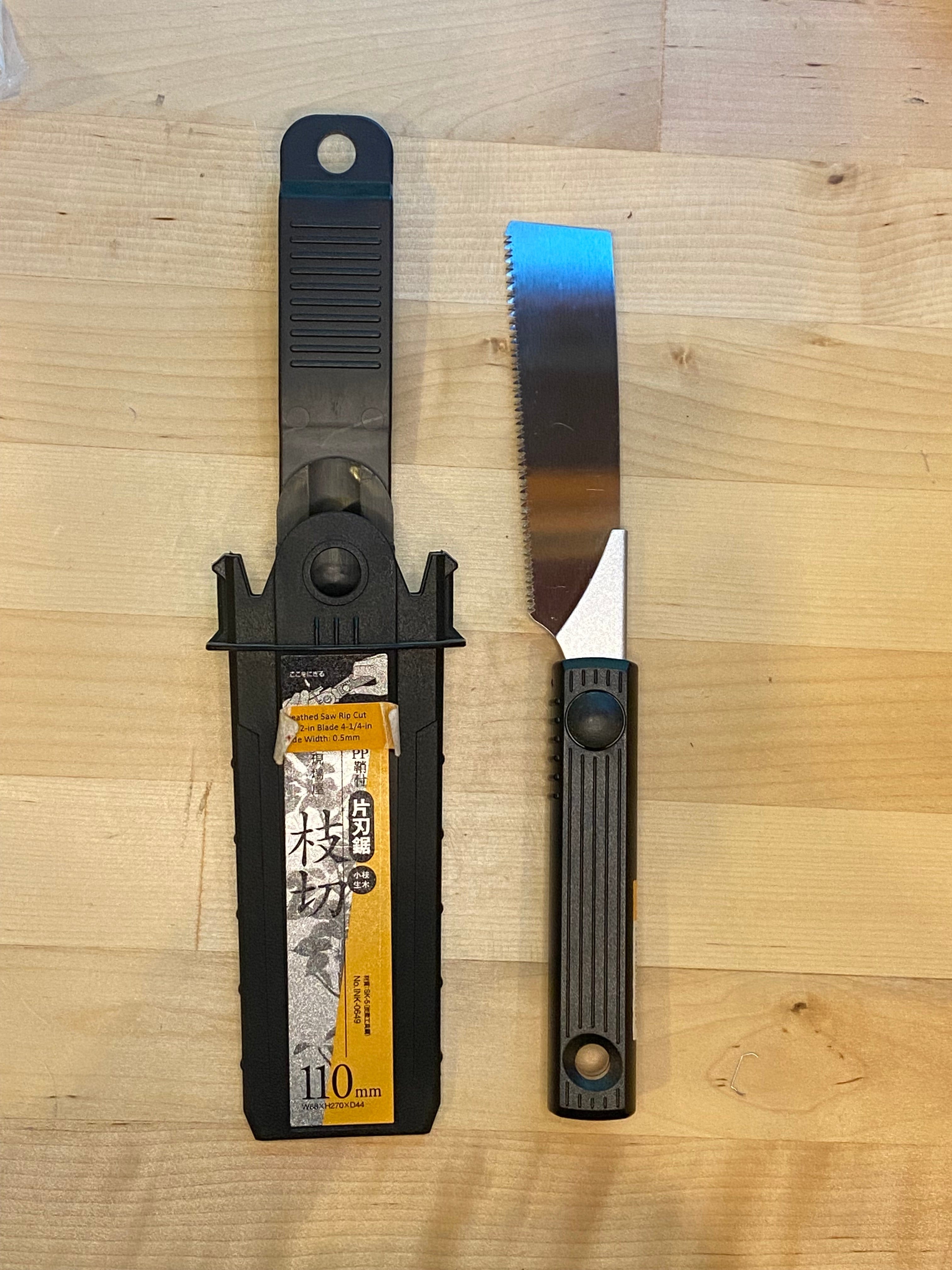 A MARU Small Sheathed Japanese Hand Saw with a black and silver handle lies next to its black plastic sheath, which features a label with text and illustrations, on a wooden surface. This fine woodworking tool boasts an impulse-hardened blade that is partially serrated.