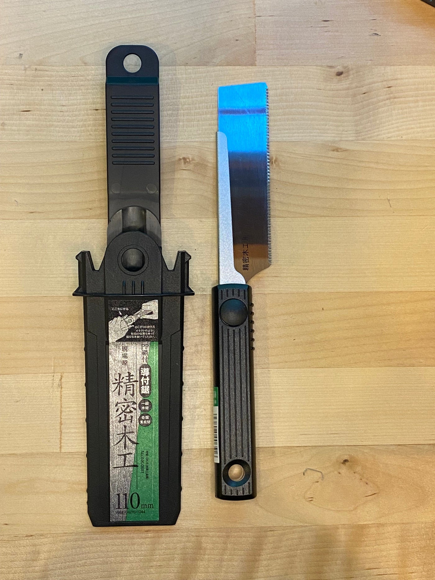 A black-handled Small Sheathed Japanese Hand Saw by MARU is placed on a light wood surface. With impulse-hardened blades and fine teeth perfect for precise cuts, the saw comes with a protective plastic sheath featuring Japanese text and a 10mm marking, making it ideal for fine woodworking.