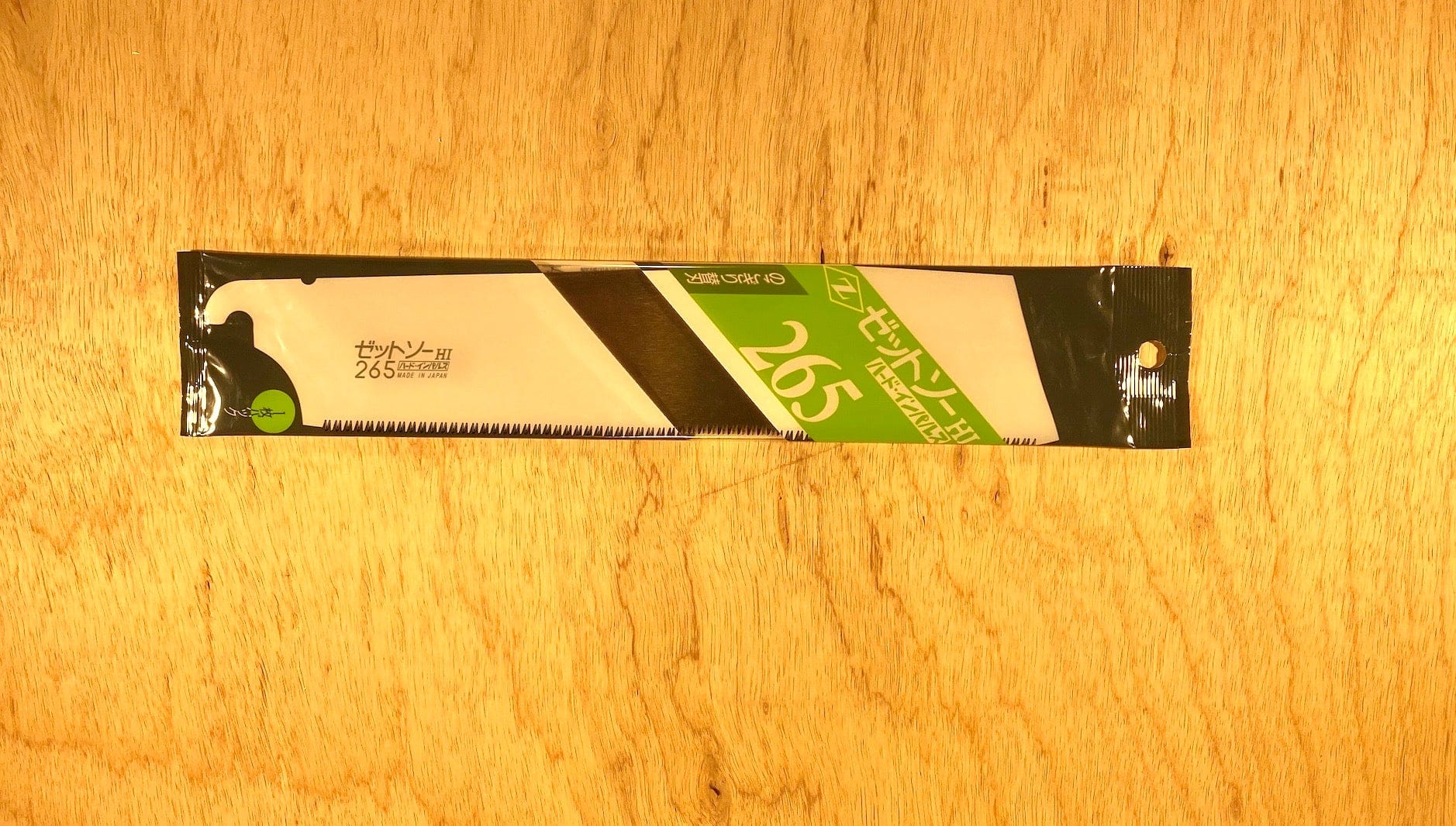 A Kataba Cross 265 by Z-Saw from MARU, in green and white packaging with Japanese text and a saw blade illustration, lies on a light wooden surface; its designed for cross cutting in carpentry applications.