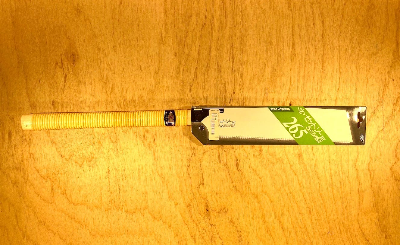 The MARU Kataba Cross 265 by Z-Saw features a yellow handle and silver blade, partially sheathed in a green and white cardboard cover on a wooden surface—perfect for precise cross-cutting in carpentry projects.