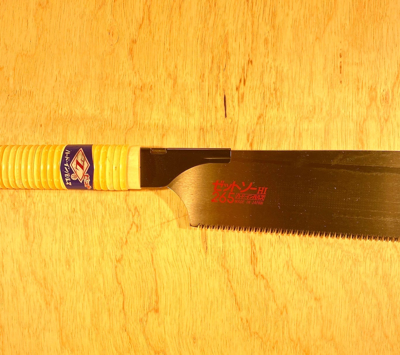 A Kataba Cross 265 by Z-Saw hand saw from MARU, with a yellow ribbed handle and black blade marked with red Japanese text and 265, rests on a light wooden surface—ideal for cross-cutting tasks.