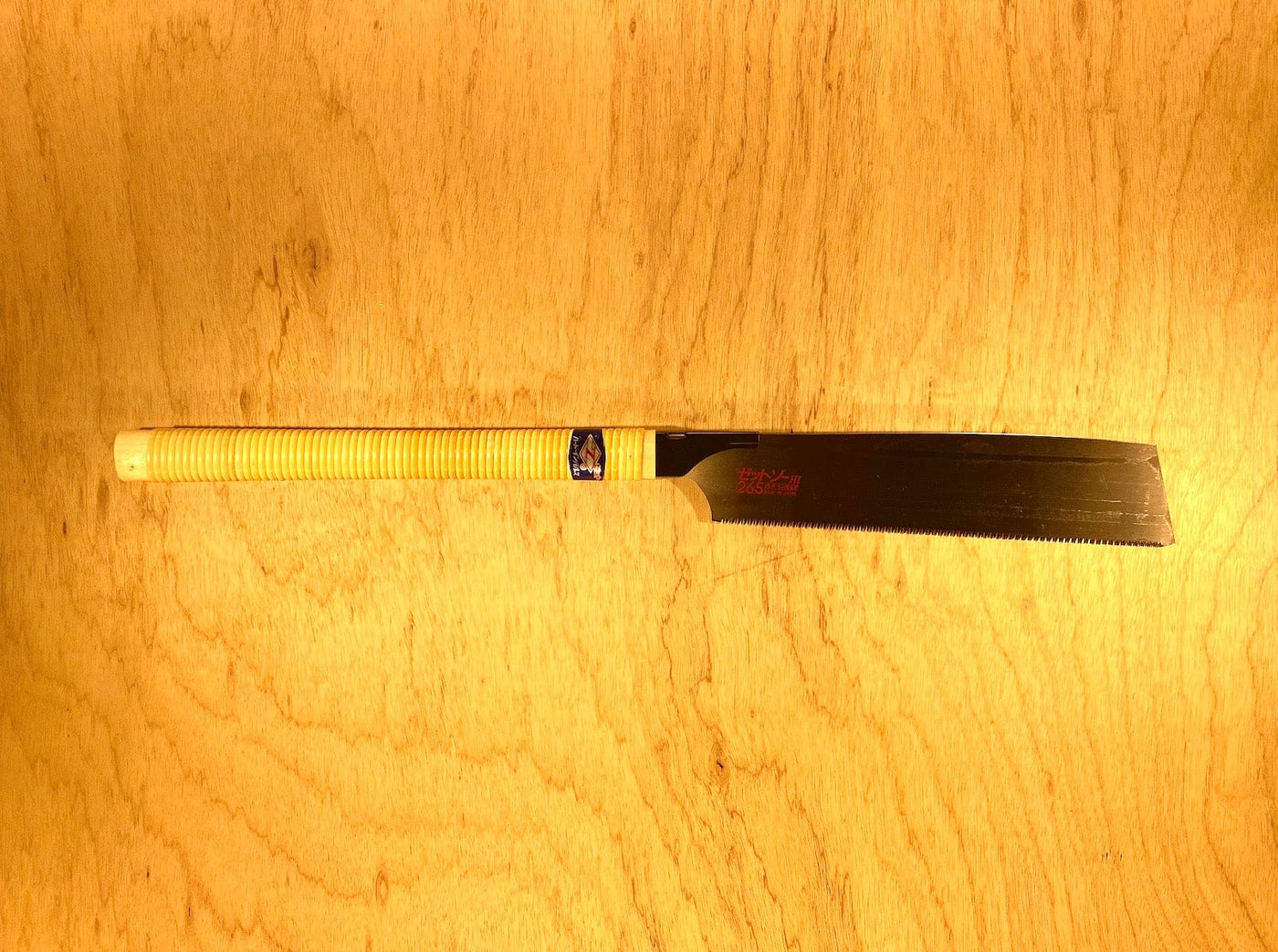 The MARU Kataba Cross 265 by Z-Saw, featuring a yellow wrapped handle and black blade, rests on a light wooden surface—perfect for precise carpentry cross cuts.