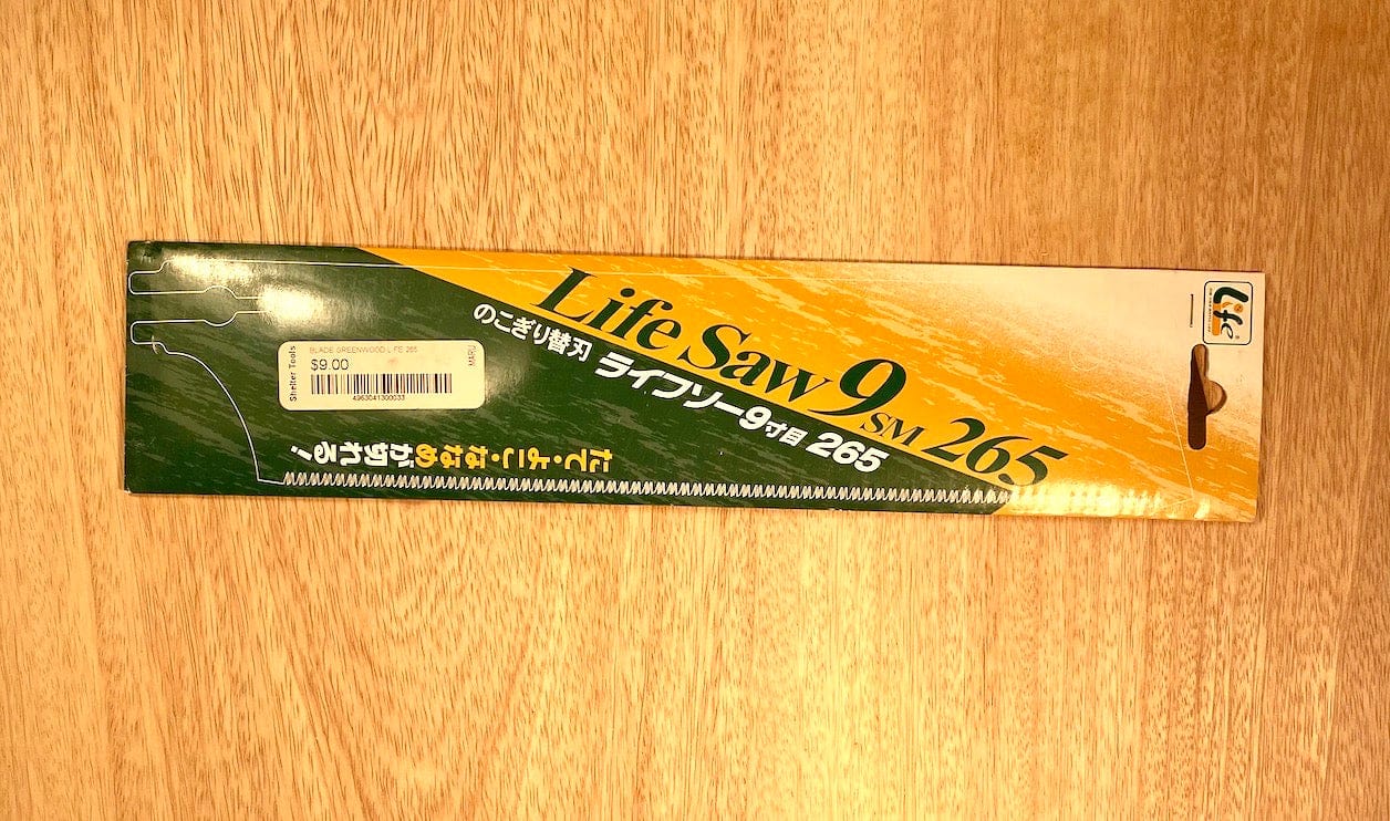 A box of Life Saw 265 by Z-Saw from the brand MARU rests on a wooden surface, displaying its green and yellow design with Japanese text. This Japanese Kataba saw includes a replaceable blade designed for efficient pull stroke cutting.