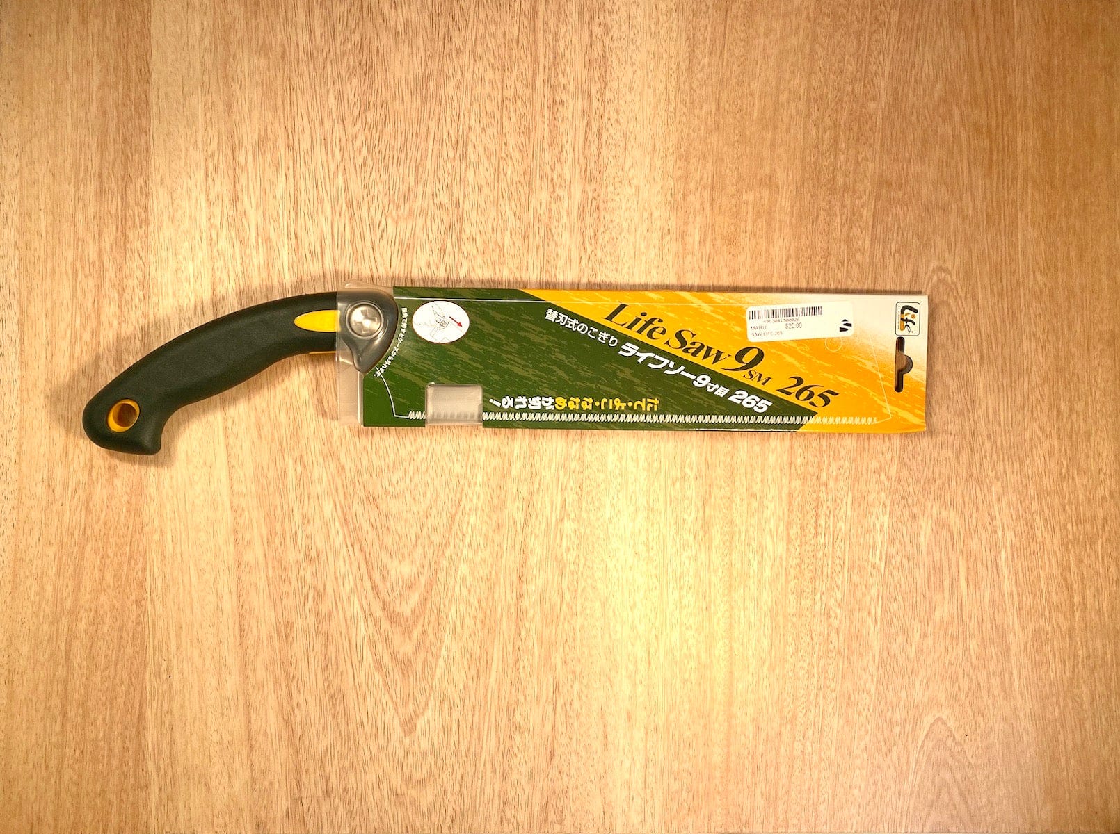 A Life Saw 265 By Z-Saw from the brand MARU, featuring a black and yellow handle, lies on a wooden surface in its cardboard sleeve. This Japanese Kataba saw is equipped with a replaceable blade and is designed for precise pull stroke cutting.