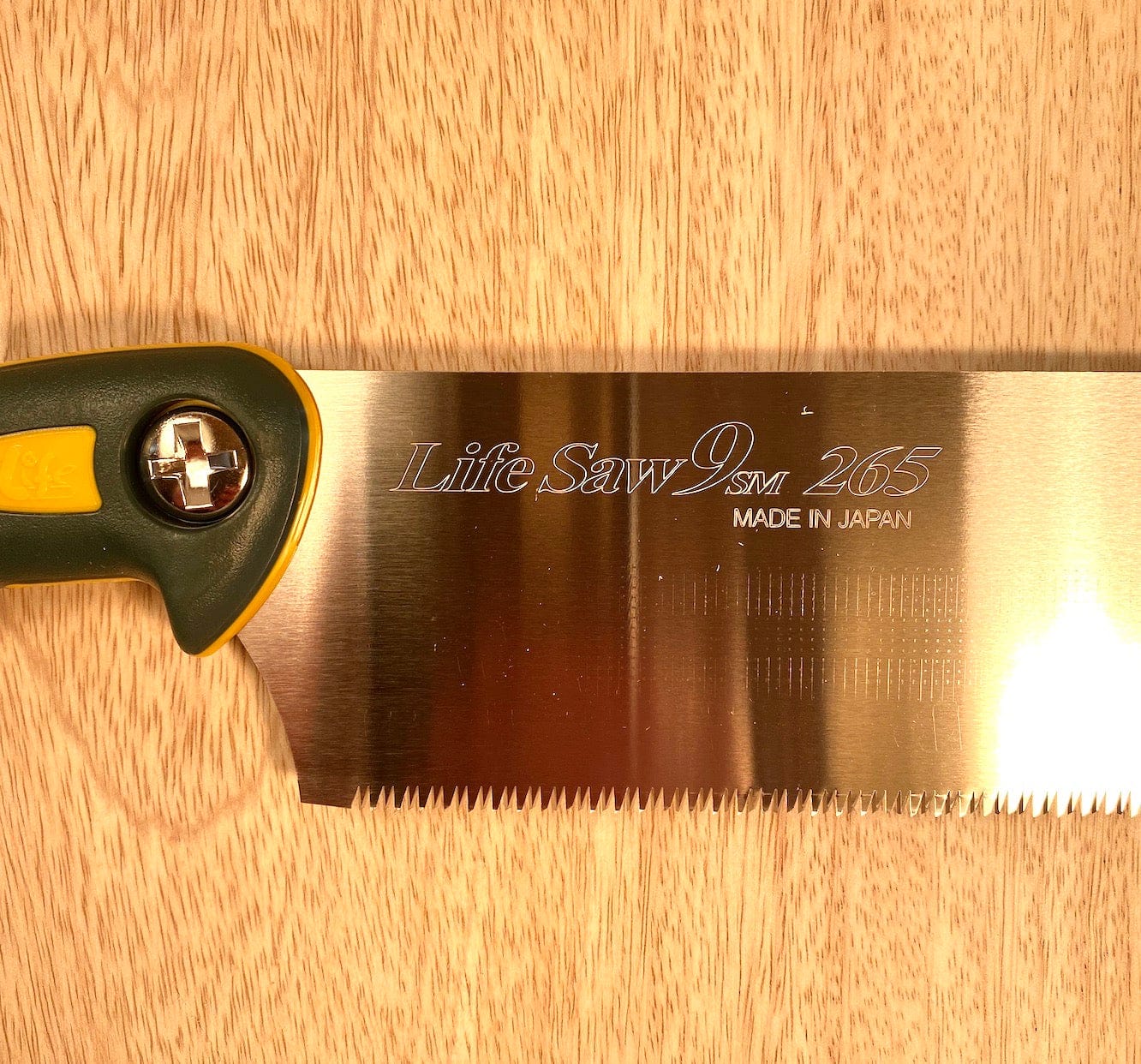 Close-up of a Life Saw 265 By Z-Saw with a shiny, replaceable blade engraved with "Made in Japan" on a light wooden surface. The handle is partially visible, featuring a green and yellow design, characteristic of a traditional Japanese Kataba Saw. The saw is branded under MARU.