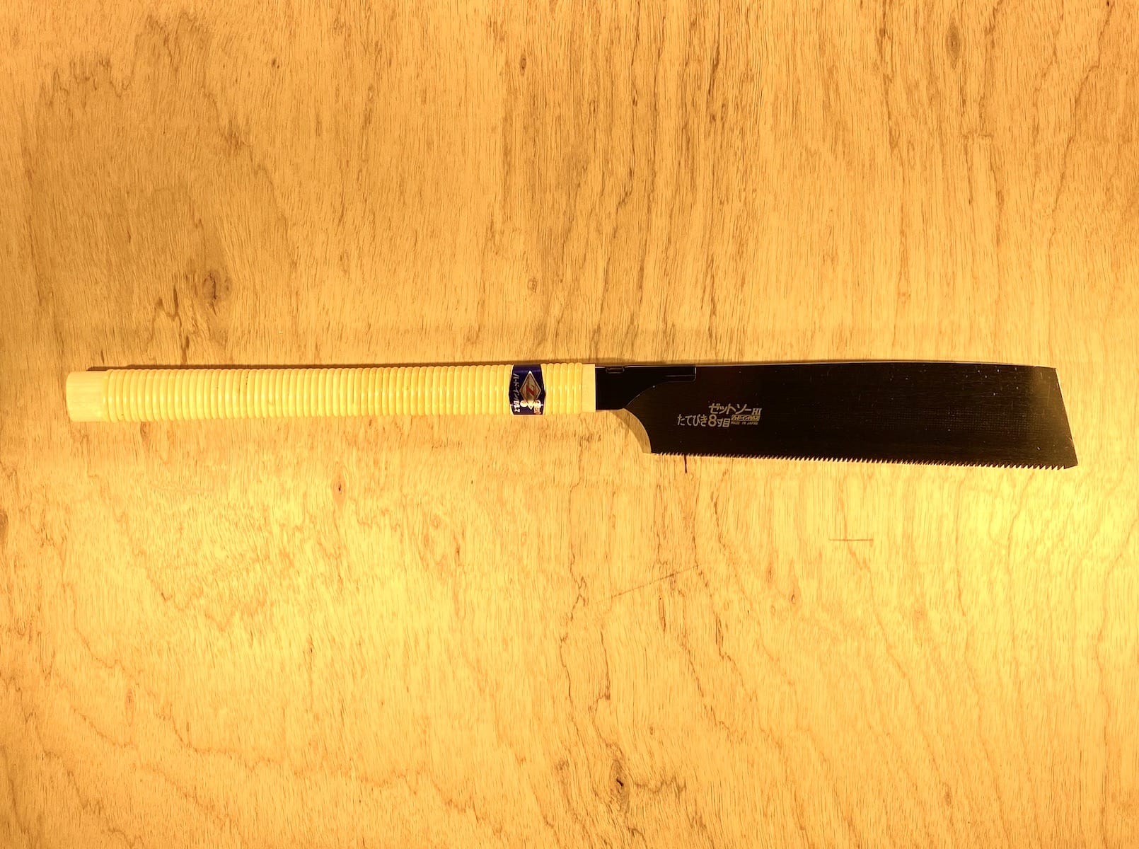 The MARU Rip H-250 by Z-Saw, featuring a black blade and white wrapped handle, lies on a light wood surface. Its classic rip-tooth-pattern teeth are visible along one blade edge.