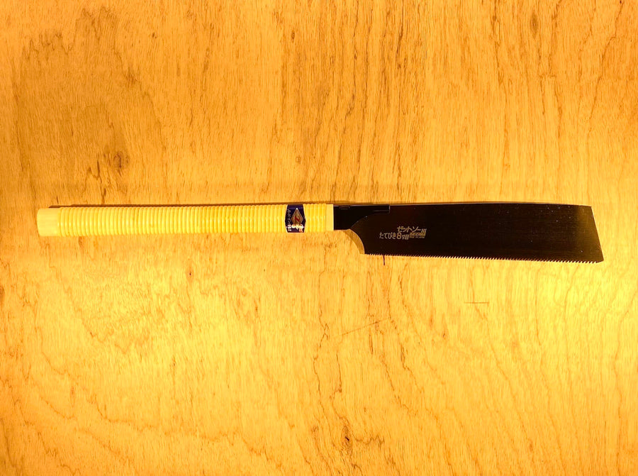 The MARU Rip H-250 by Z-Saw, featuring a black blade and white wrapped handle, lies on a light wood surface. Its classic rip-tooth-pattern teeth are visible along one blade edge.